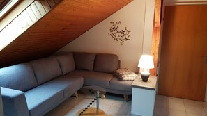 Quarto de canto com sofá cinzento, uma mesinha com abajur, um adorno na parede e uma porta de madeira.
