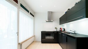 A modern kitchen with black cabinetry, a stainless steel range hood, and a window with blinds. - Appartements Hofquartier (Lucerne)
