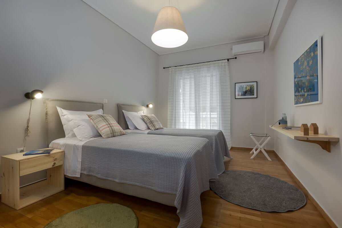 Une chambre à coucher comprenant un lit, des tables de chevet, une lampe suspendue, une fenêtre avec des rideaux, une petite étagère et un tableau accroché au mur.