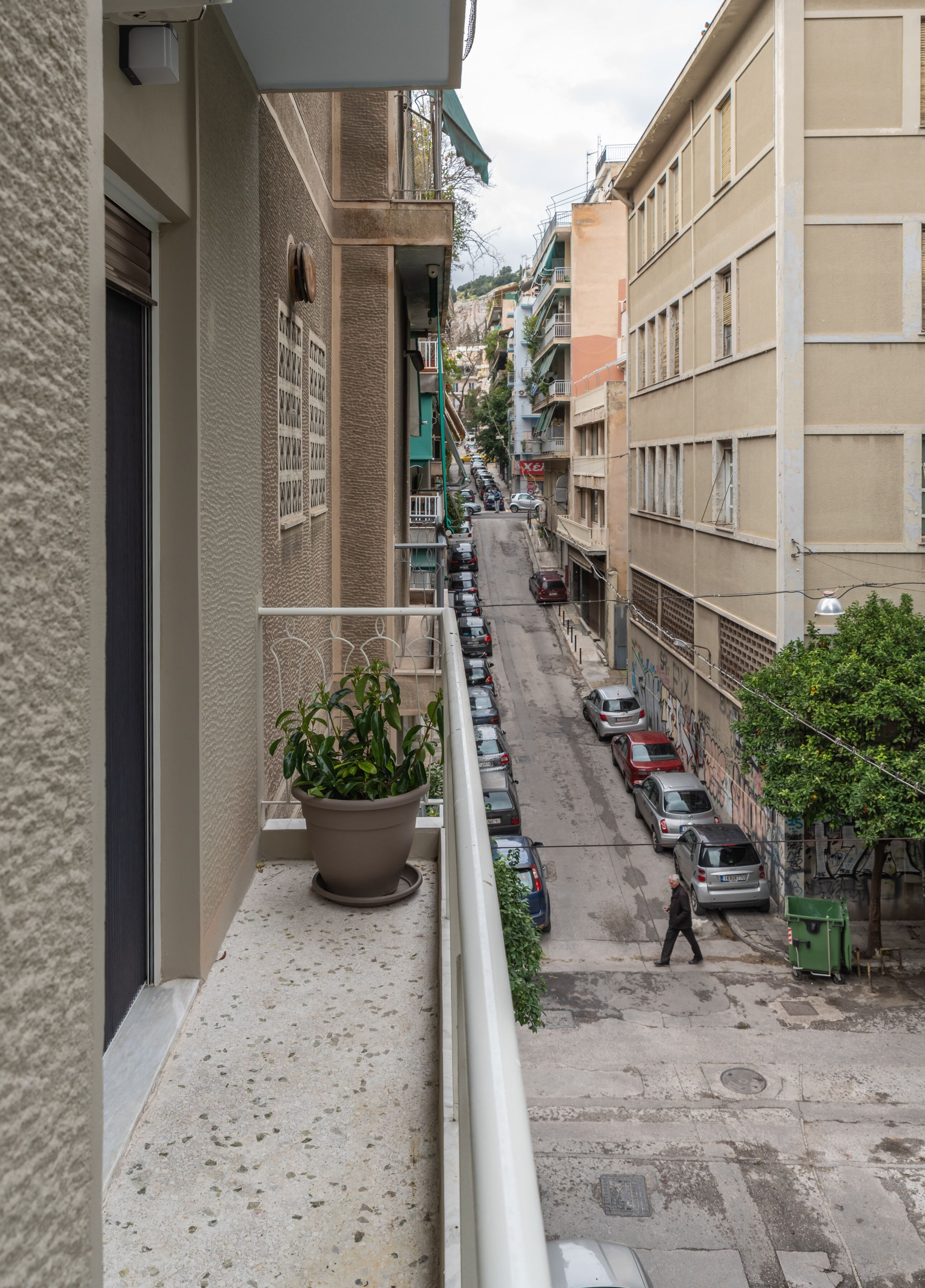 A narrow street with parked cars, a balcony with a potted plant, and buildings with balconies and windows.