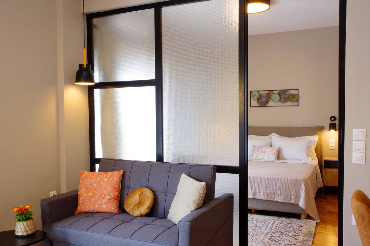 Une chambre moderne avec un canapé gris, une petite table d’appoint sur laquelle est posé un vase de fleurs, et un lit avec une tête de lit.
