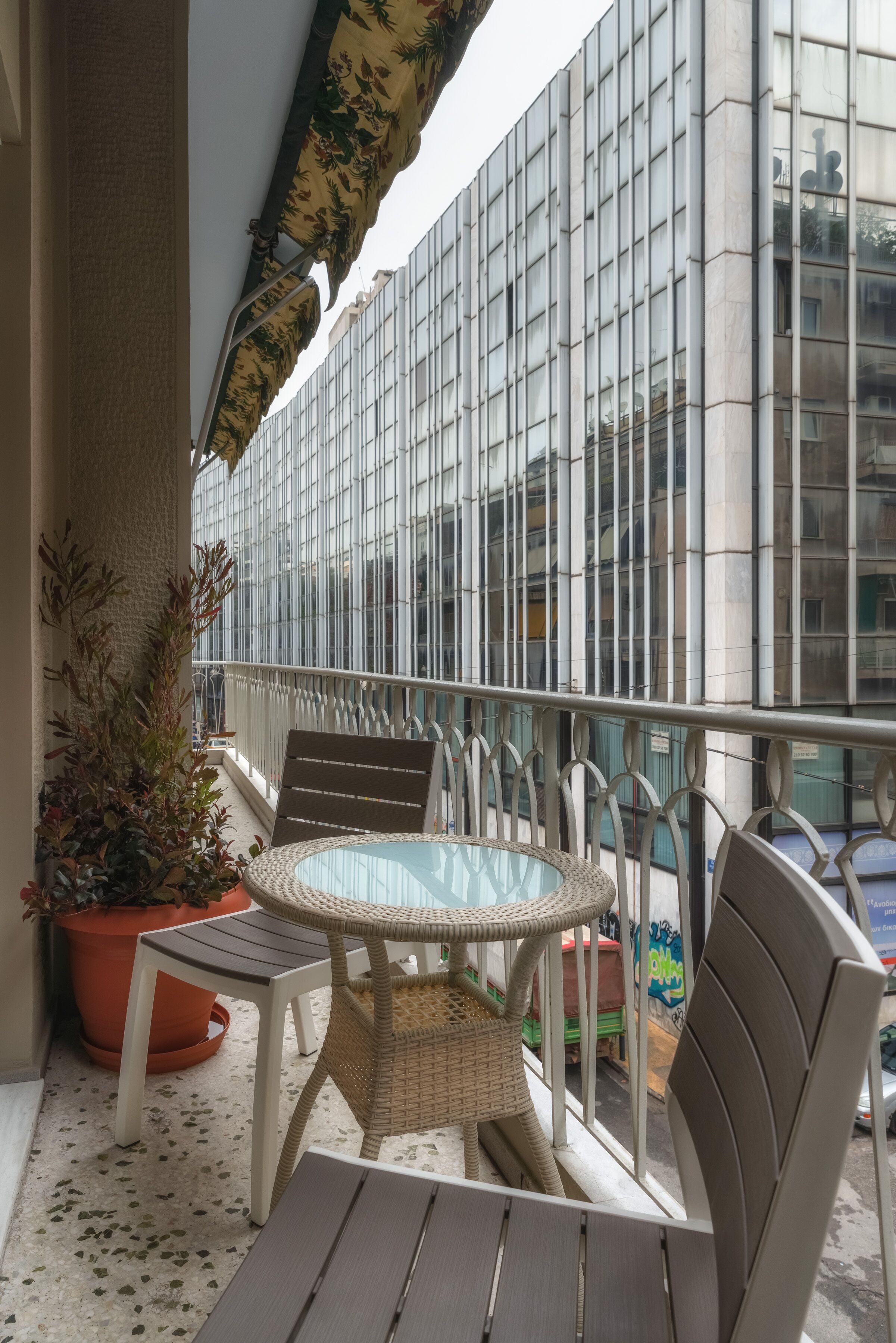 Uma varanda com mesa e cadeiras de vidro, um vaso de planta e vista para um edifício com janelas amplas.
