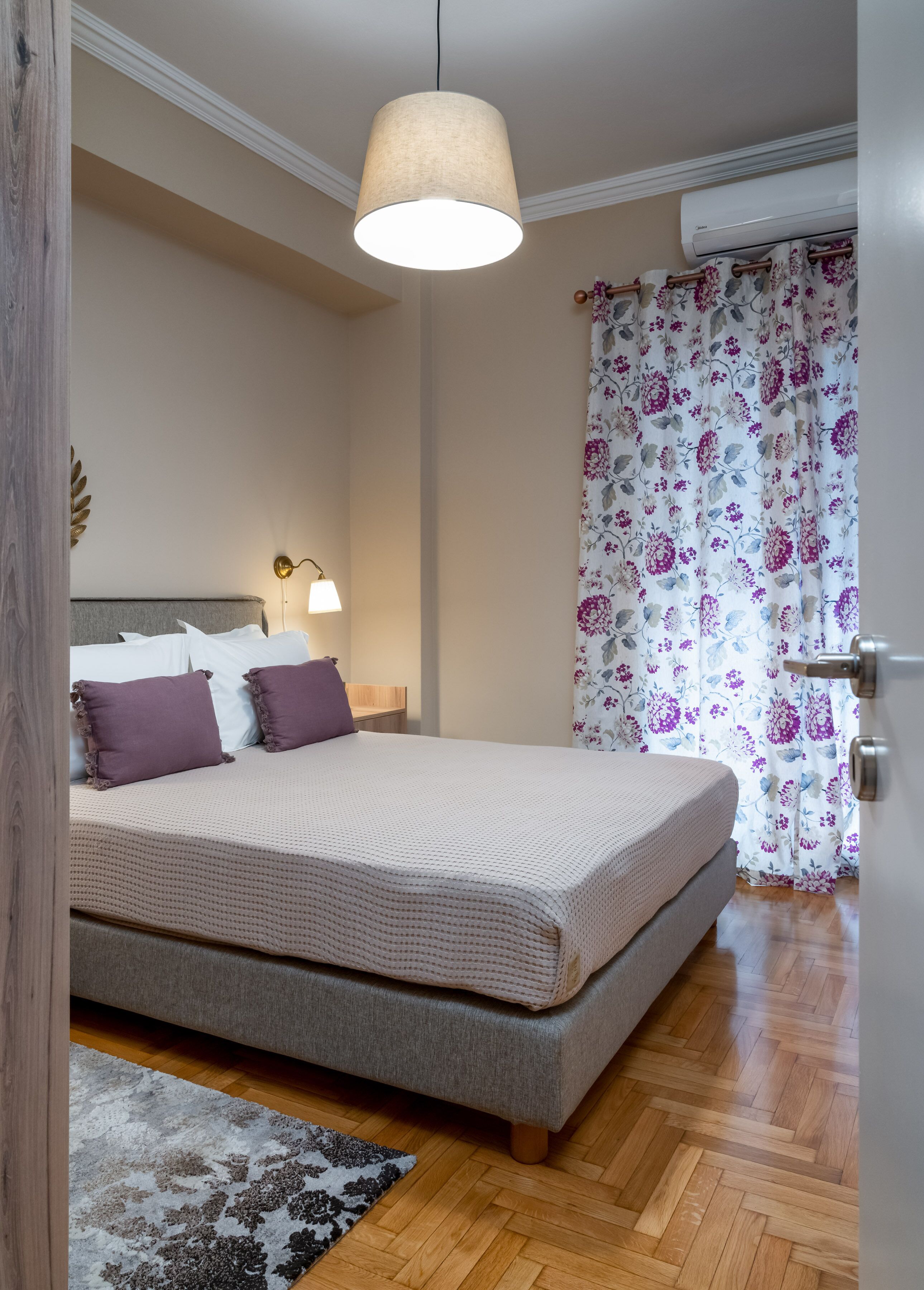 A bedroom with a bed, a bedside lamp, a floral curtain, and a wooden floor.
