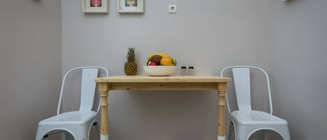 A room with a checkered floor, a wooden table, two white chairs, a fruit bowl, and two framed pictures on the wall.
