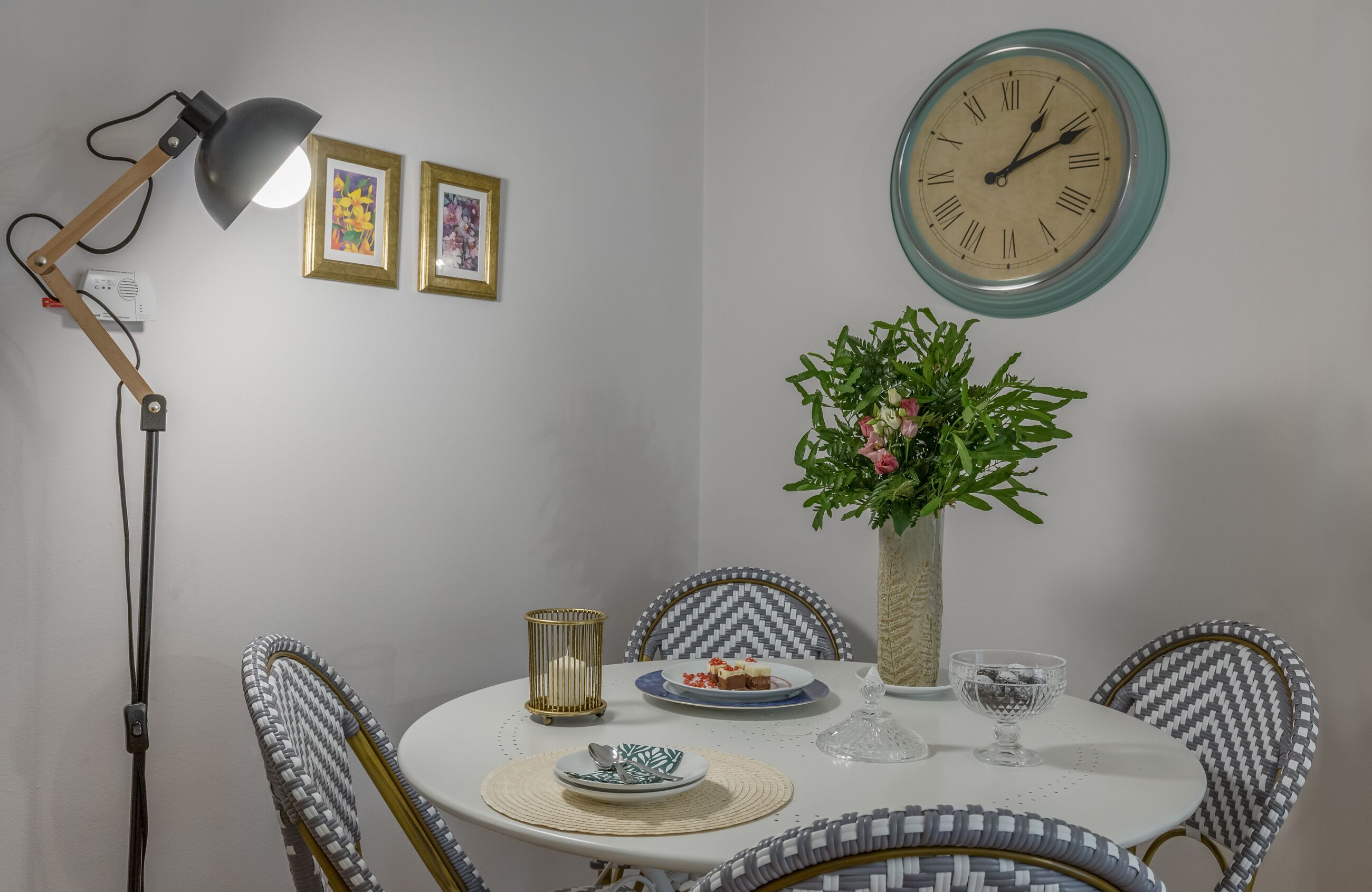 A dining area with a round table, chairs, a wall clock, a lamp, framed pictures, and a potted plant.