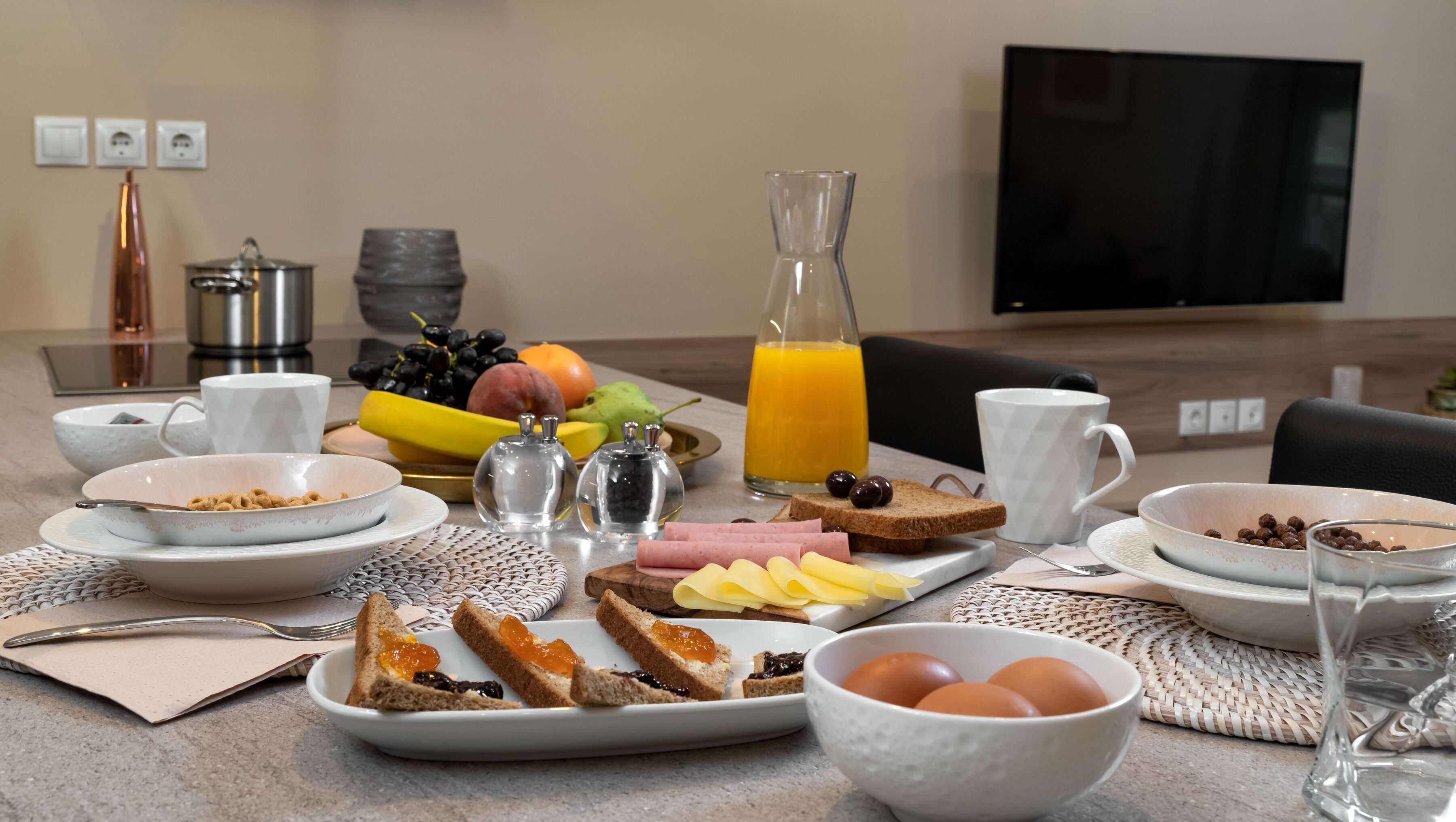A breakfast table with various food items, a glass of orange juice, and a TV in the background.
