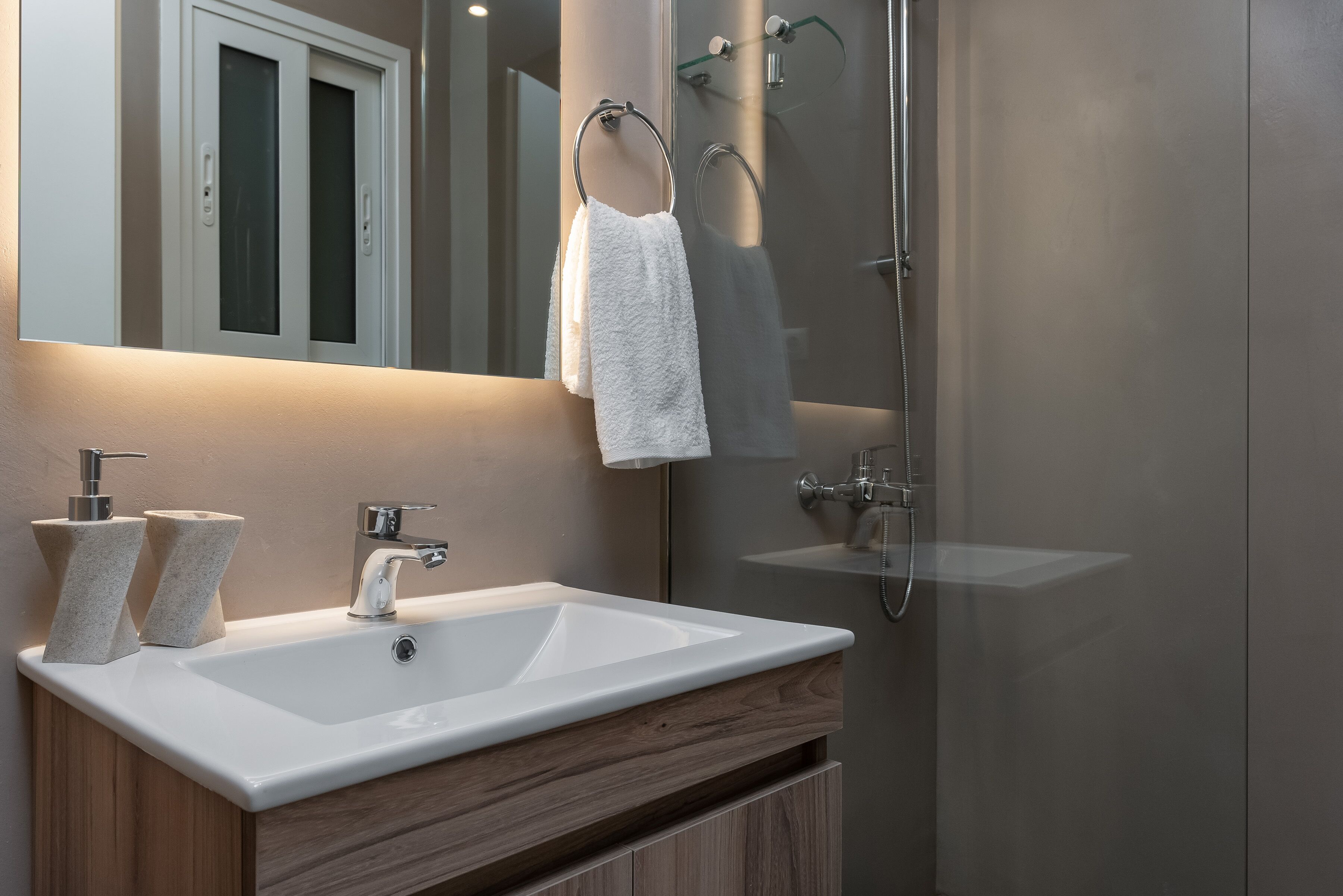 A modern bathroom with a white sink, two towel dispensers, a mirror, and a glass-enclosed shower.