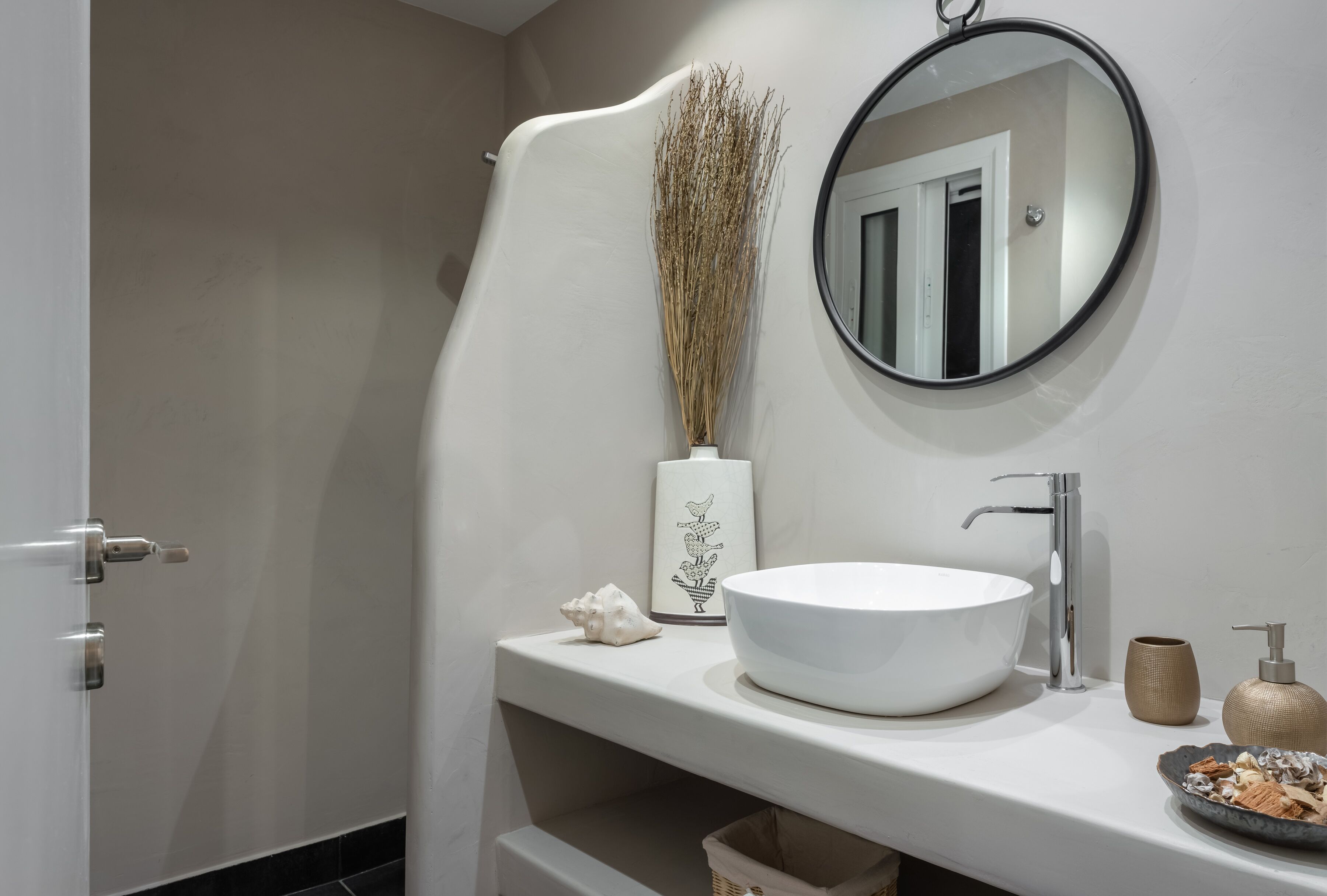 A modern bathroom with a white sink, a round mirror, and a vase with dried plants.