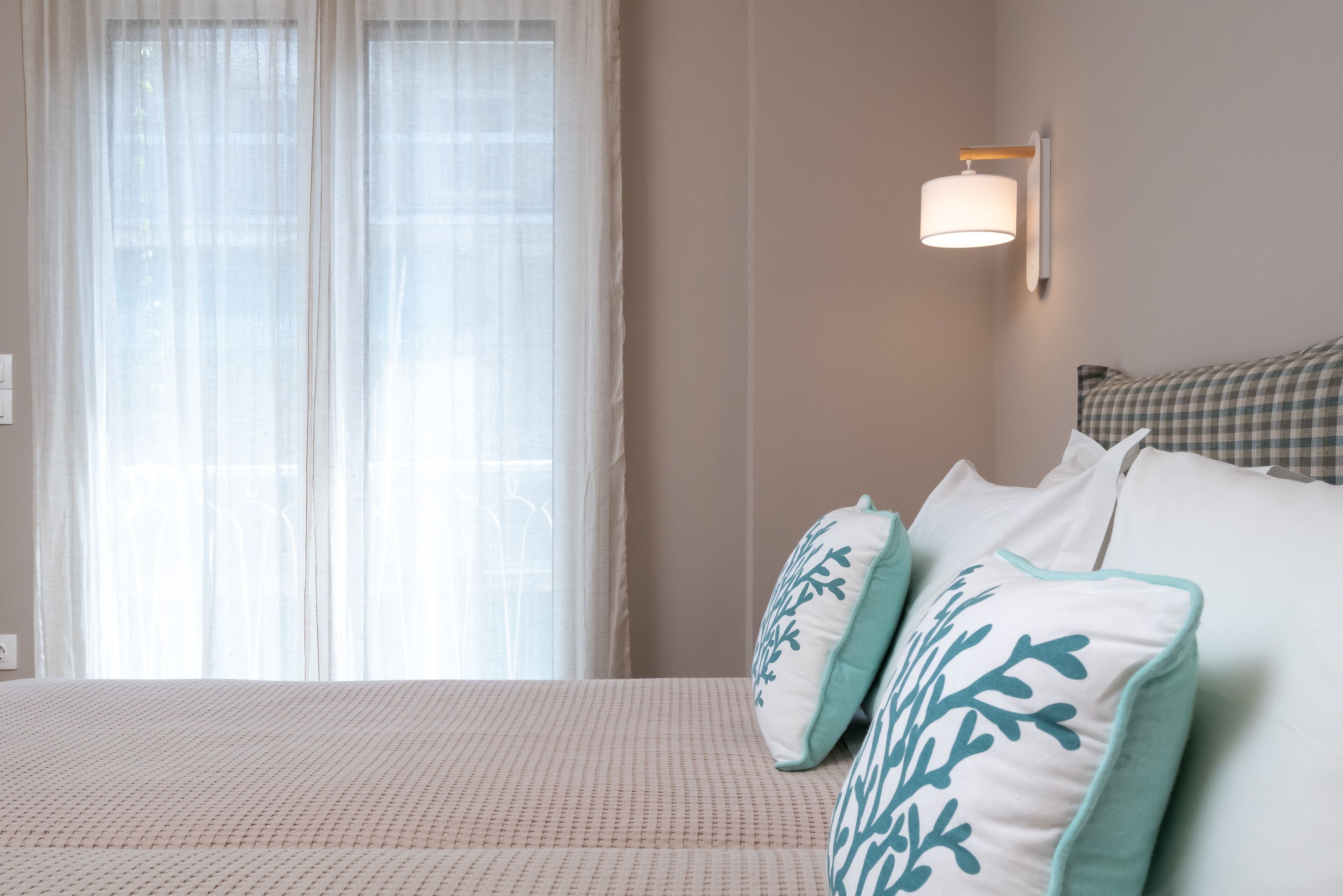 A bedroom with a bed, two pillows with a leaf pattern, a wall-mounted light, and sheer curtains.