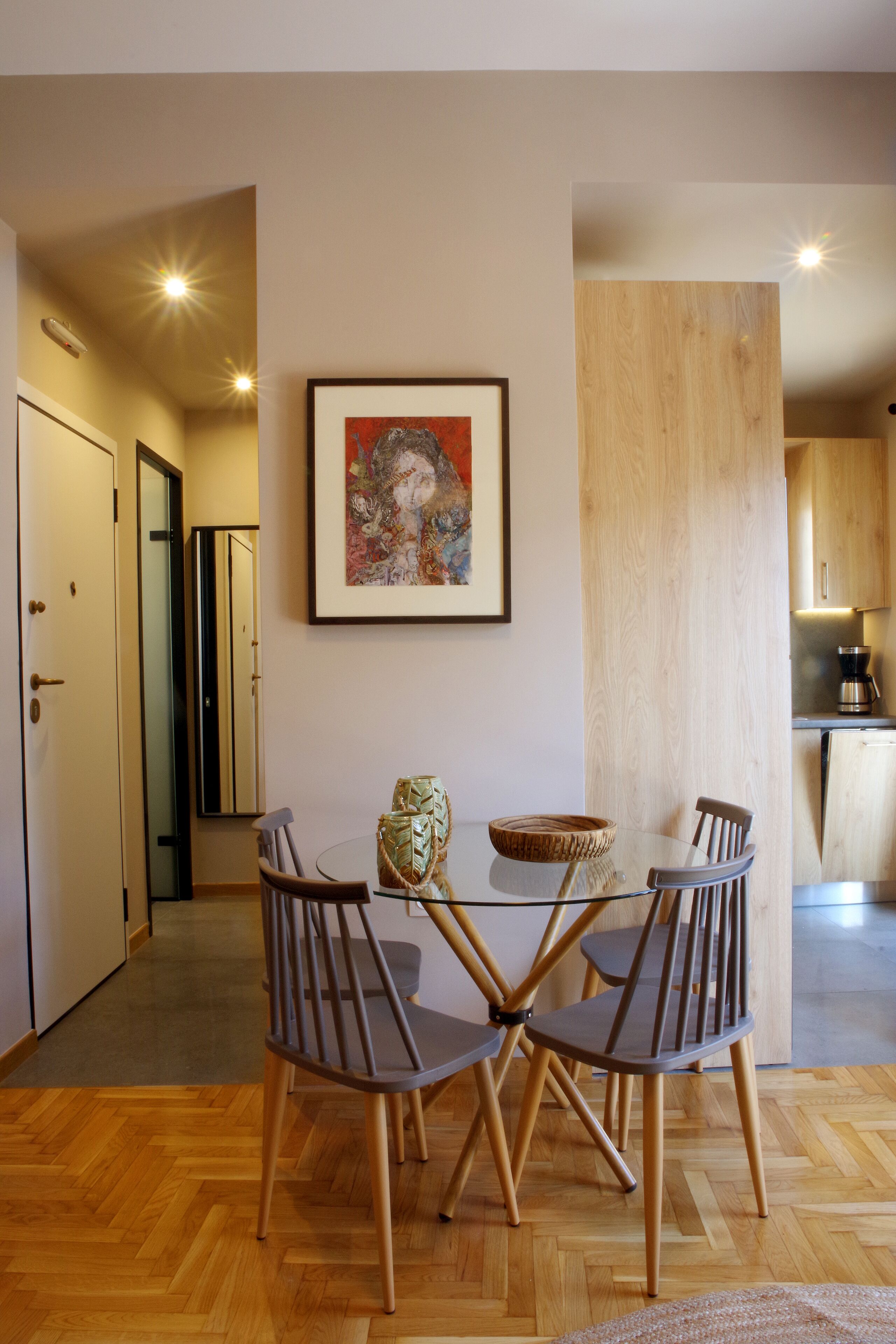 A dining area with a glass table and four chairs, a framed artwork on the wall, and a wooden cabinet in the background.