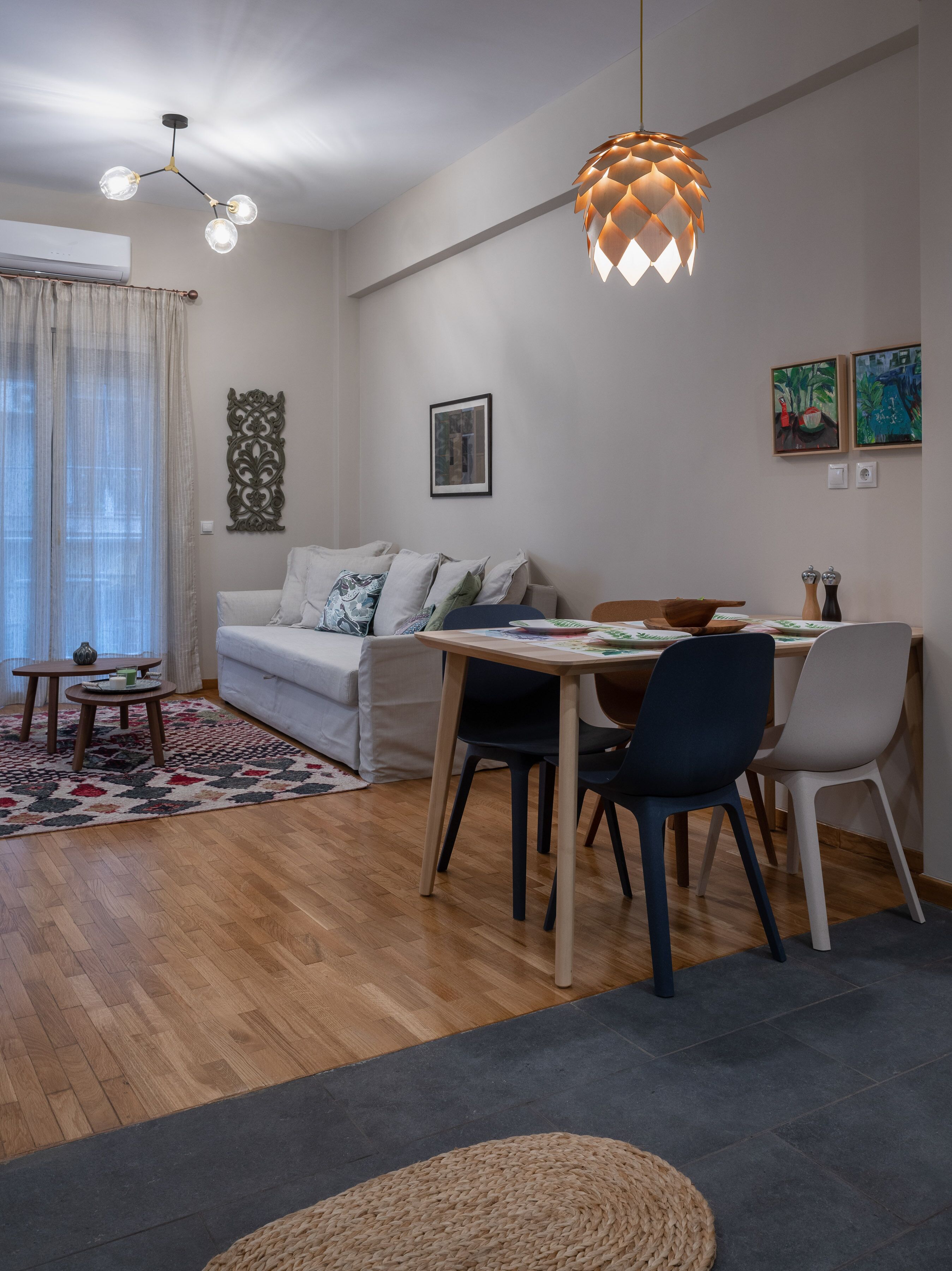 A modern living room with a dining area, featuring a wooden floor, a white sofa, a dining table with chairs, a hanging pendant light, and artwork on the walls.