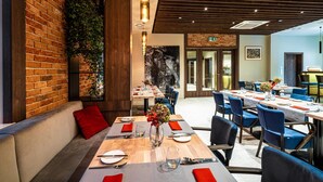 Un restaurant aux murs de briques, au plafond en bois et aux chaises bleues. Les tables sont dressées avec des serviettes rouges et des couverts en argent.