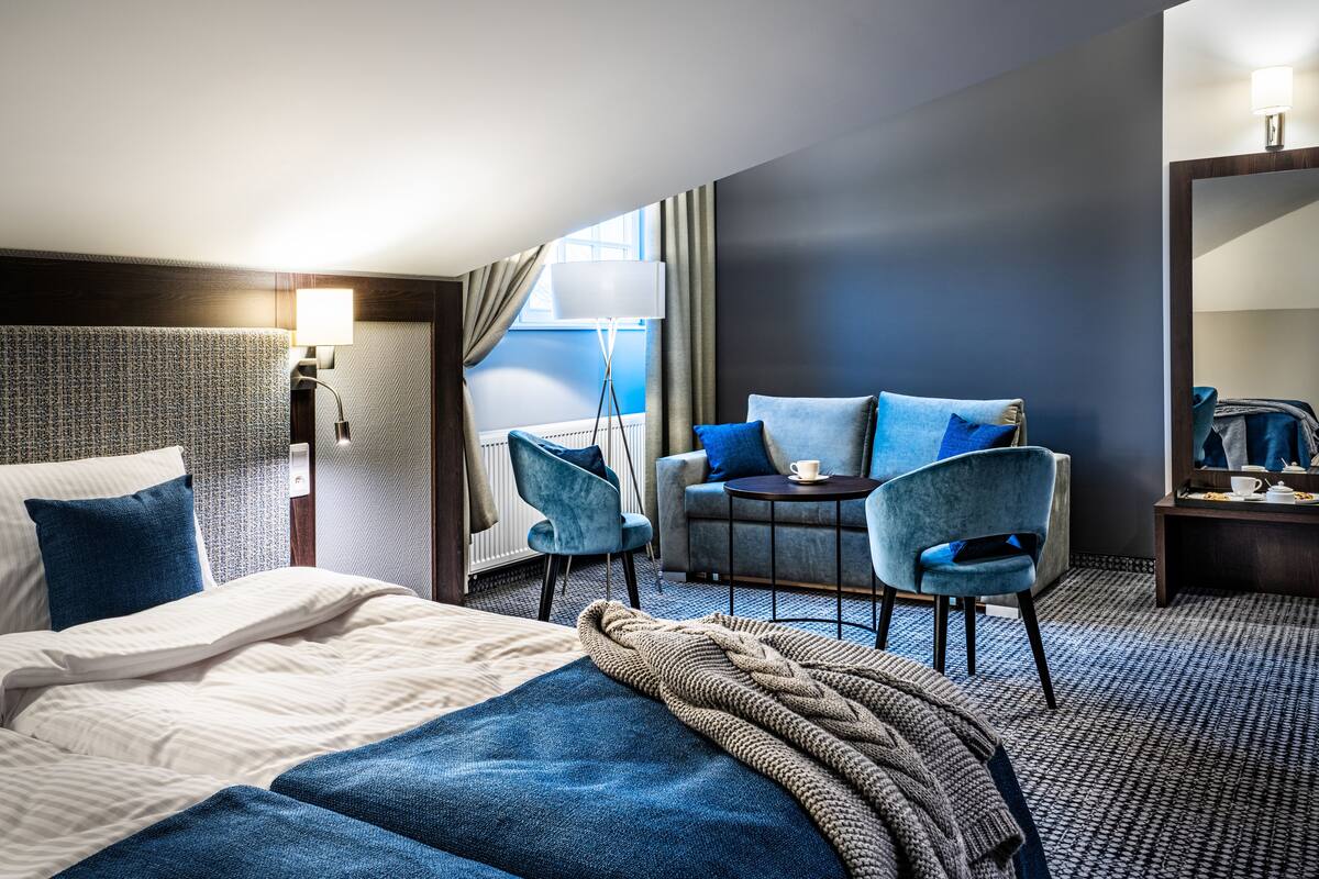 Une chambre d’hôtel avec un lit, un bureau, deux chaises, une lampe et une fenêtre avec des rideaux.