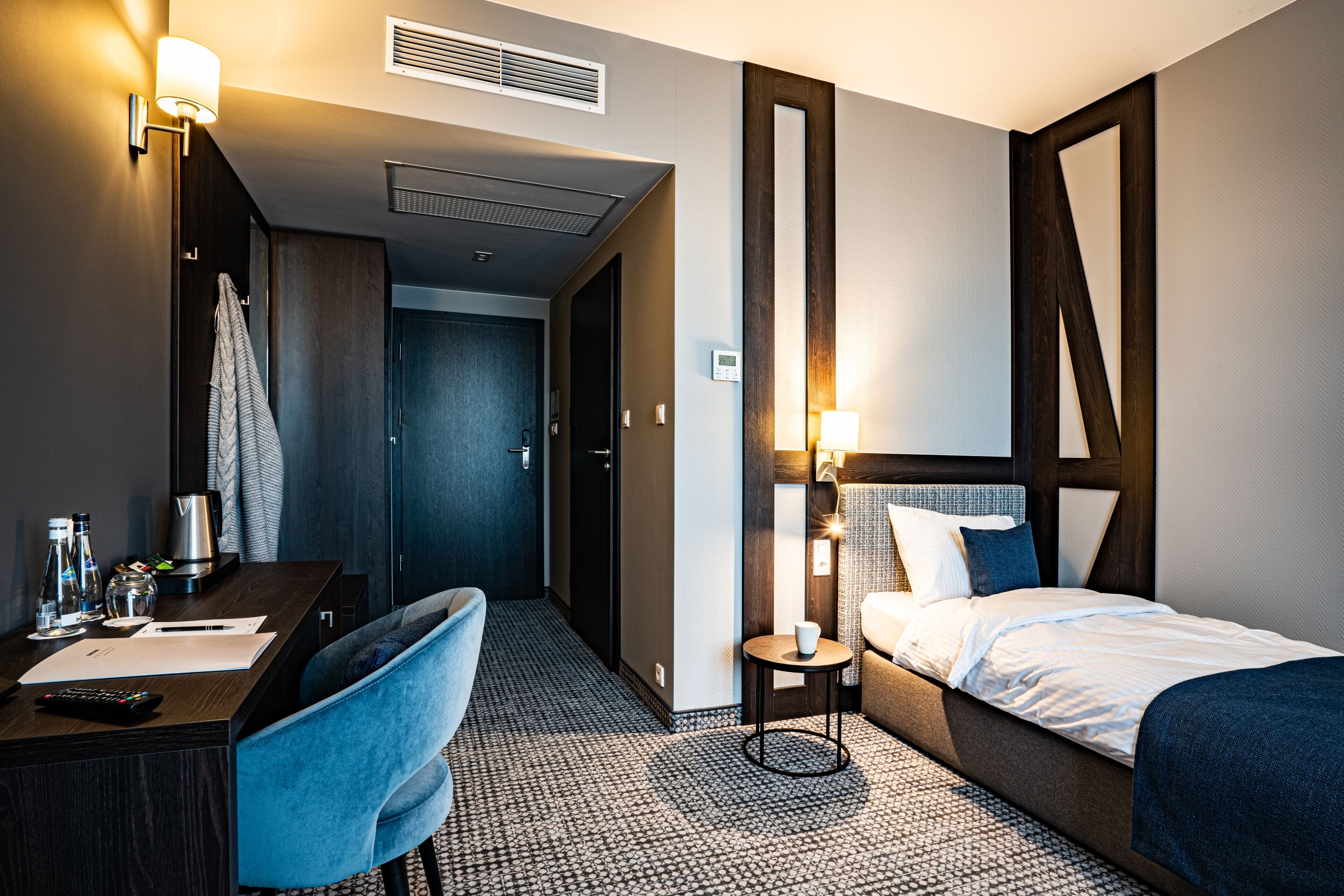 Ein modernes Hotelzimmer mit Bett, Schreibtisch mit Stuhl, einem kleinen Tisch mit Lampe und einem durch eine offene Tür einsehbaren Badezimmer.