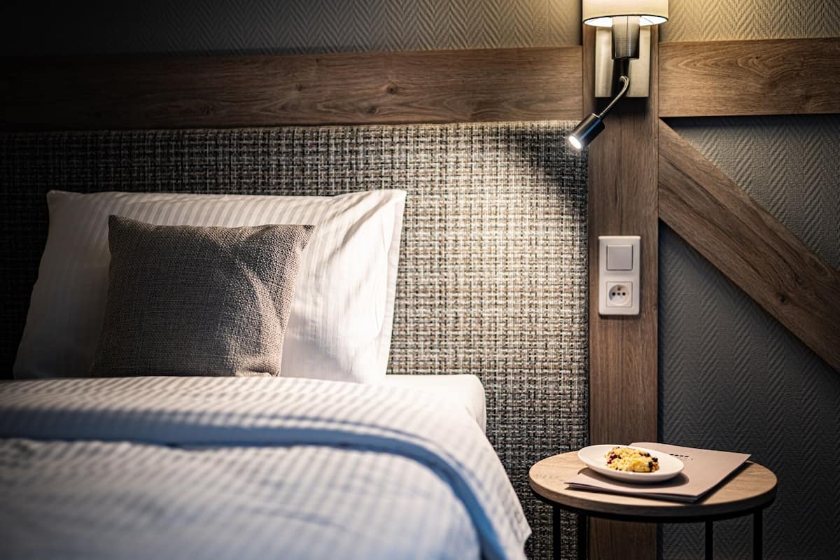 Un lit bien fait, doté d’une tête de lit texturée, une table de chevet ronde sur laquelle se trouve une assiette de nourriture, et un luminaire fixé au mur.