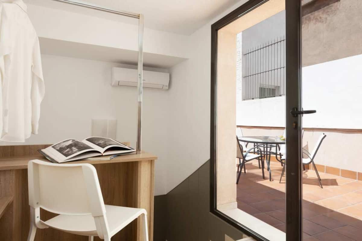 Una habitación moderna con un escritorio de madera, una silla blanca y un ventanal con vistas a un balcón con mesa y sillas.