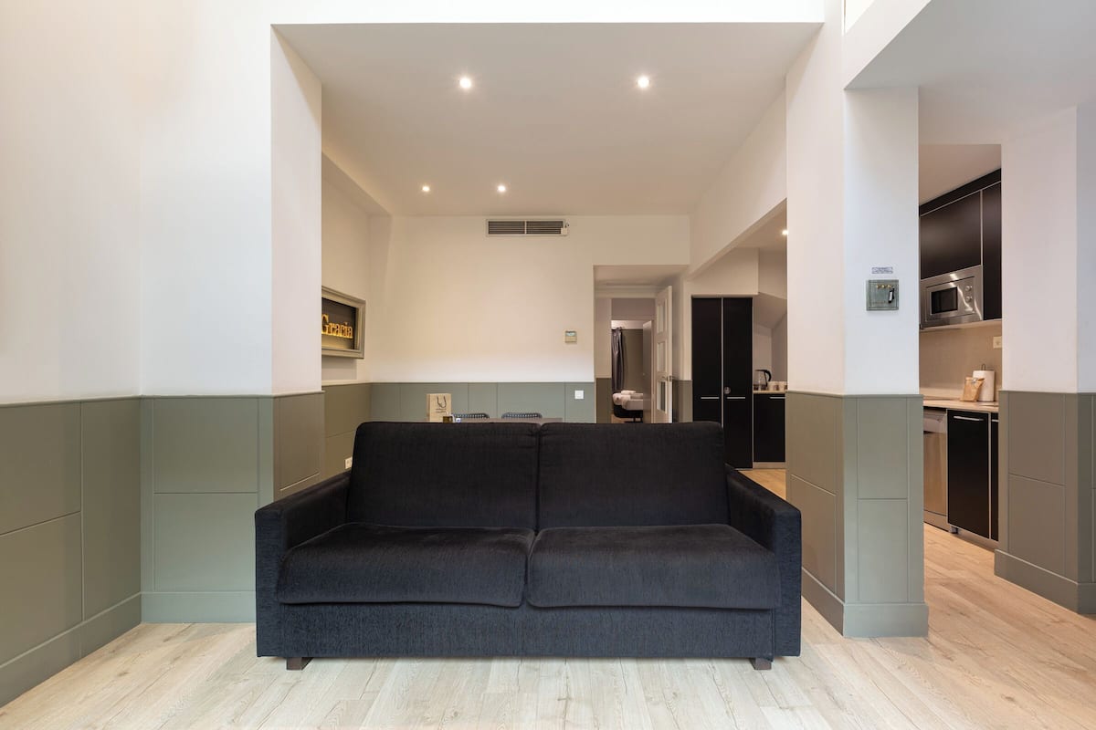 Una sala de estar moderna con un sofá de color oscuro, una cocina con electrodomésticos de acero inoxidable y una paleta de colores neutros.