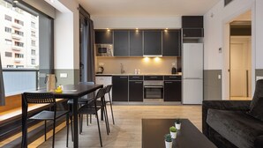 Un apartamento moderno con cocina, comedor y sala de estar. Cuenta con muebles de cocina oscuros, una mesa de comedor con sillas, una mesa de centro con plantas y un amplio ventanal con vista a un edificio.