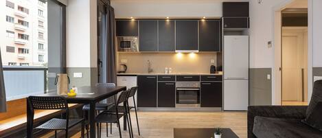 A modern apartment with a kitchen, dining area, and living room. It features dark cabinetry, a dining table with chairs, a coffee table with plants, and a large window offering a view of a building.