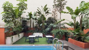 Zona de piscina al aire libre con borde de azulejos, rodeada de abundante vegetación y un espacio de descanso con mesas y sillas.