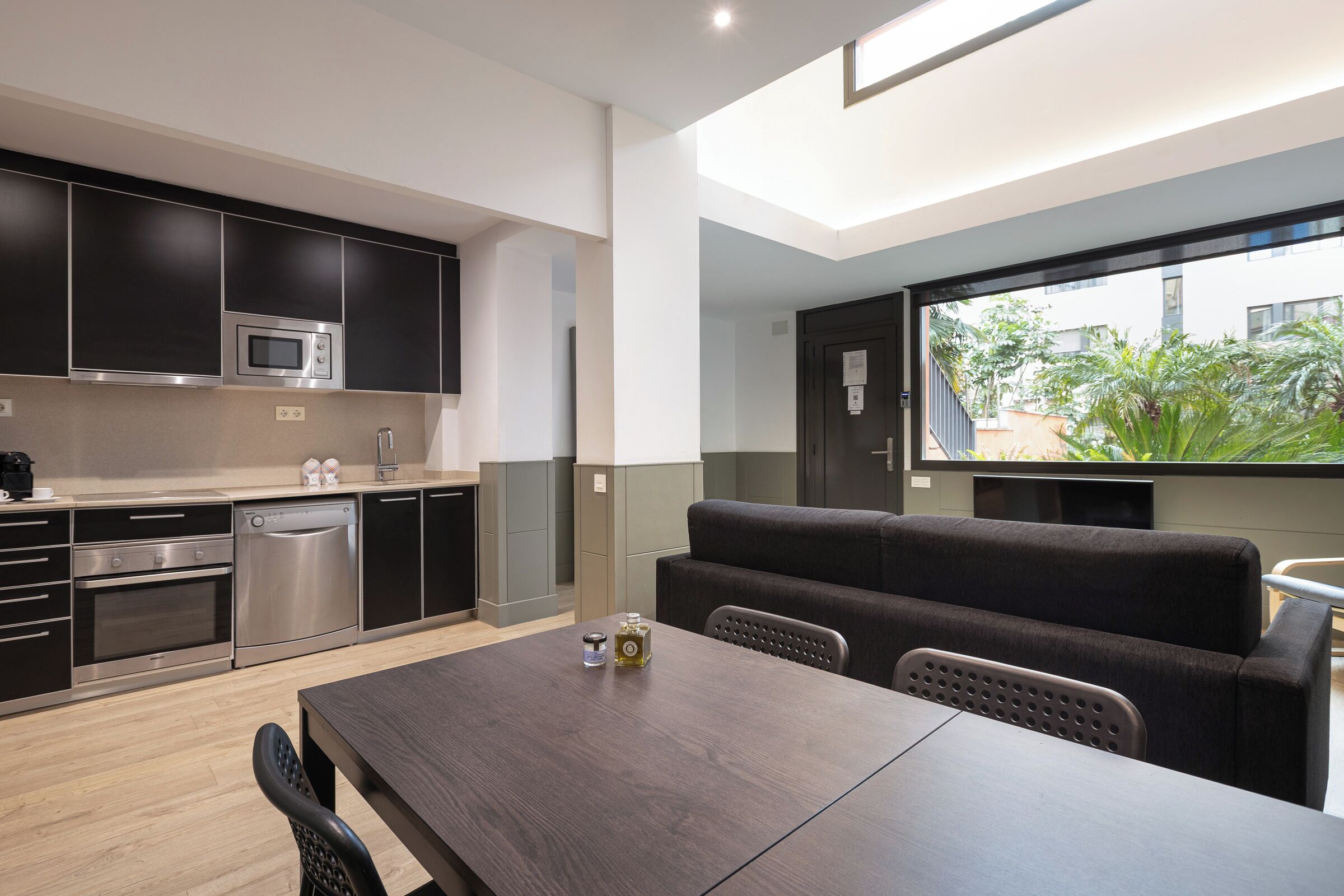 Une cuisine moderne avec des meubles de couleur sombre, des appareils en acier inoxydable et un coin repas comprenant une table en bois et des chaises noires.