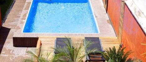 Una piscina rectangular con revestimiento azul, rodeada por una terraza de madera y plantas de palmera.