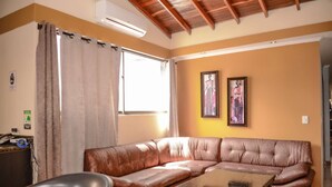 Una sala de estar con un sofá de cuero marrón, un sillón rojo y una mesita. Hay una ventana con cortinas, un equipo de aire acondicionado y dos cuadros en la pared.