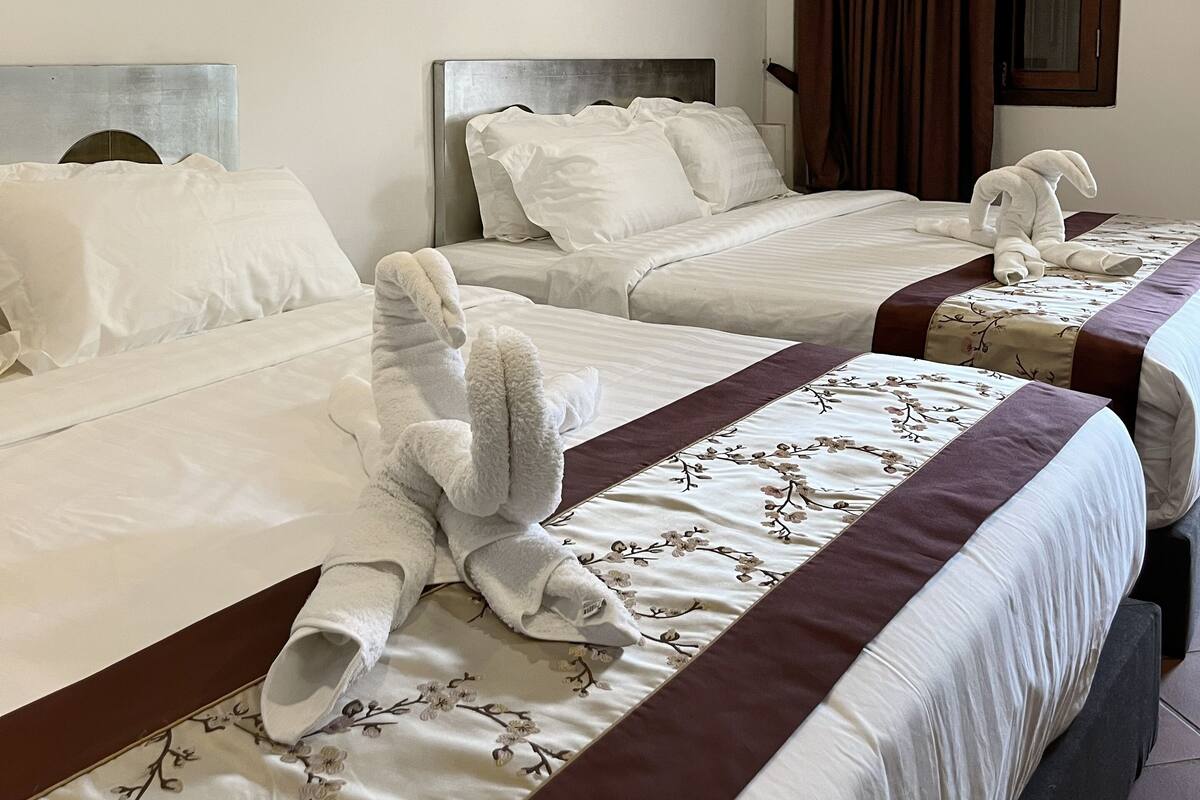 Ein Hotelzimmer mit zwei Betten, jeweils mit weißer Bettwäsche und dekorativen Kissen. An der Wand hängt ein Gemälde und ein Fenster mit Vorhängen.