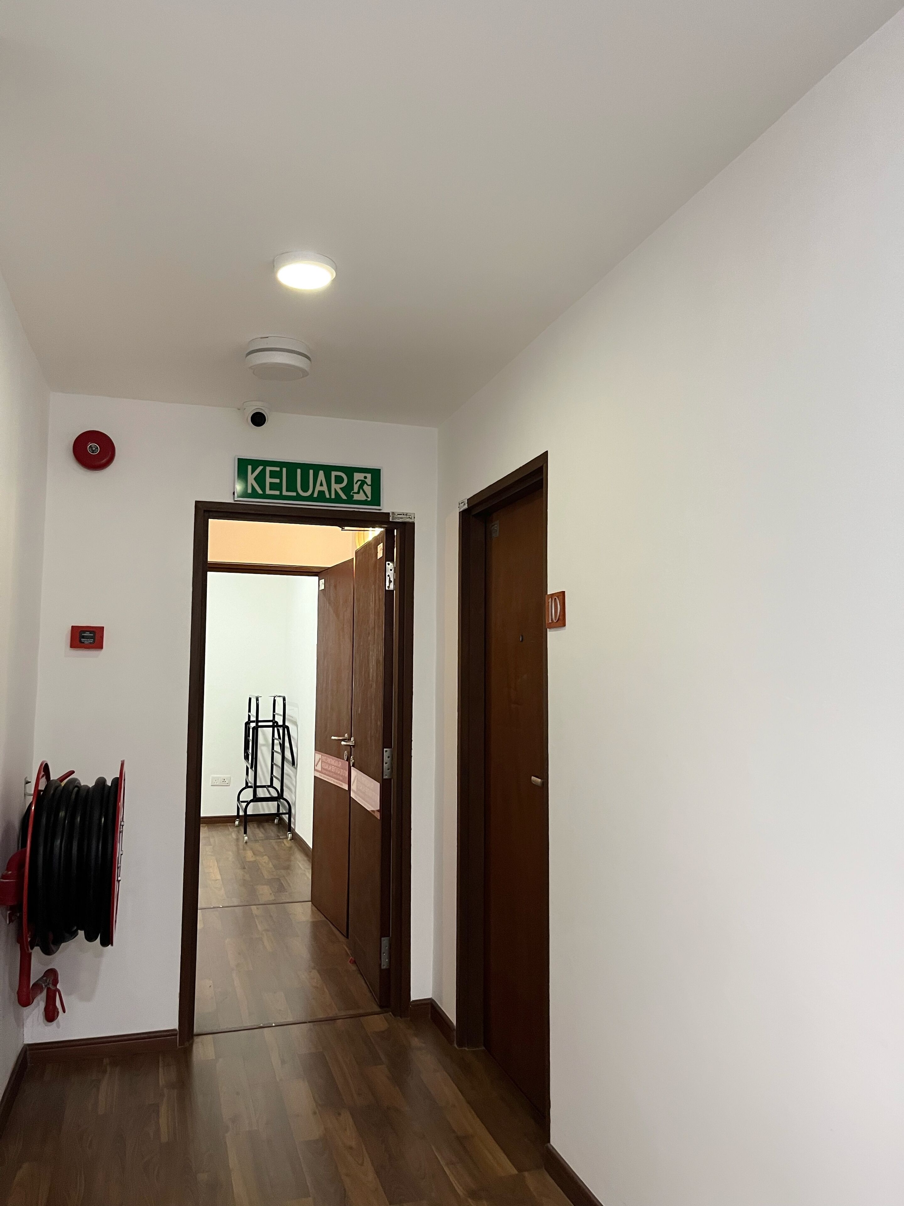 Un pasillo con un letrero que dice “KELUAR” que da a una habitación. Hay un perchero con objetos encima, una puerta con una placa de número y un detector de incendios en la pared.