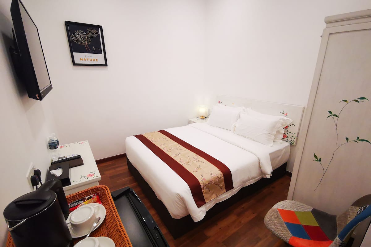 Ein ordentlich eingerichtetes Hotelzimmer mit Bett, Nachttisch mit Lampe, kleinem Tisch mit Kaffeemaschine und Stuhl mit farbigem Kissen.