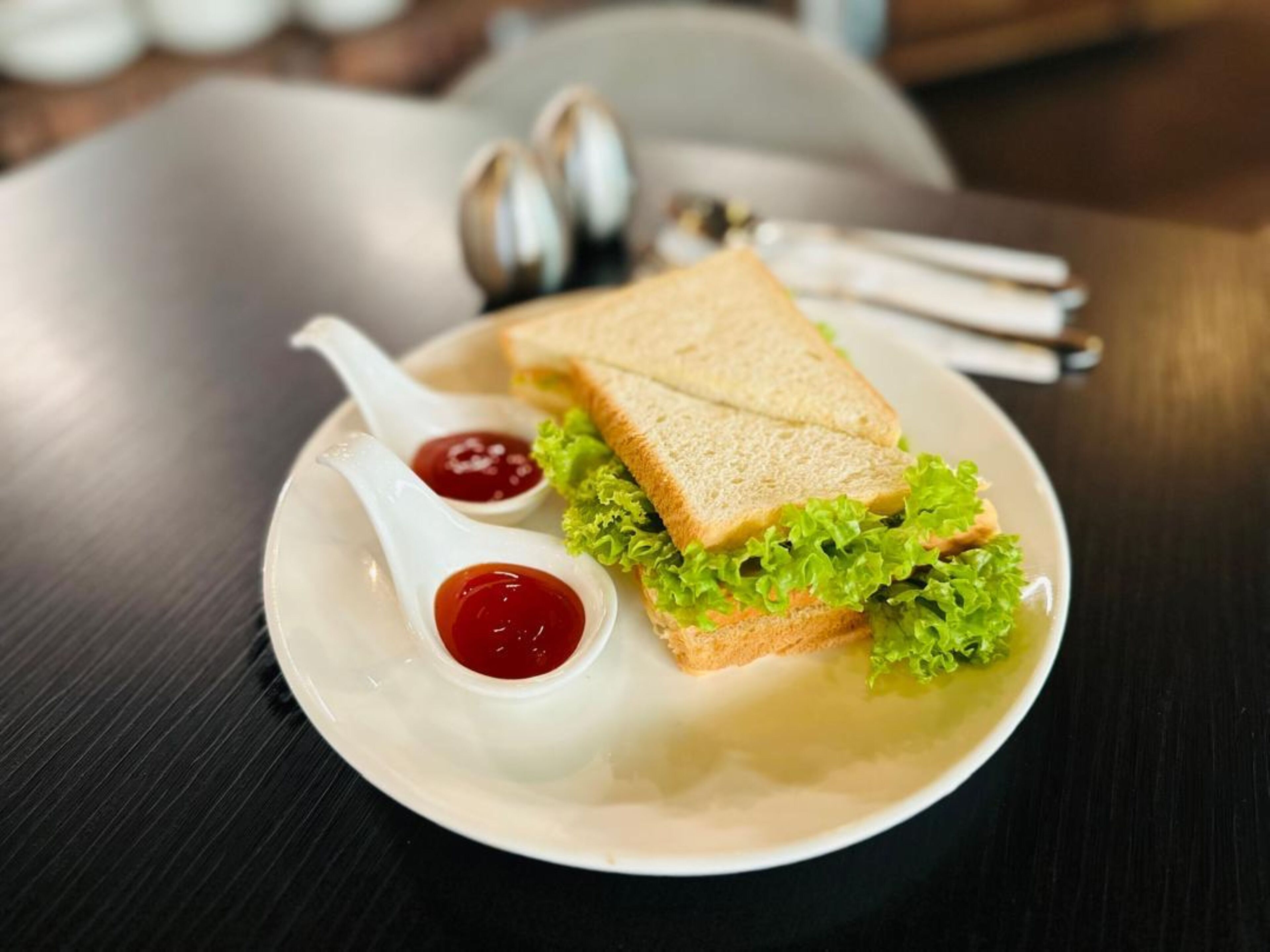 A sandwich on a plate with two spoons of ketchup.