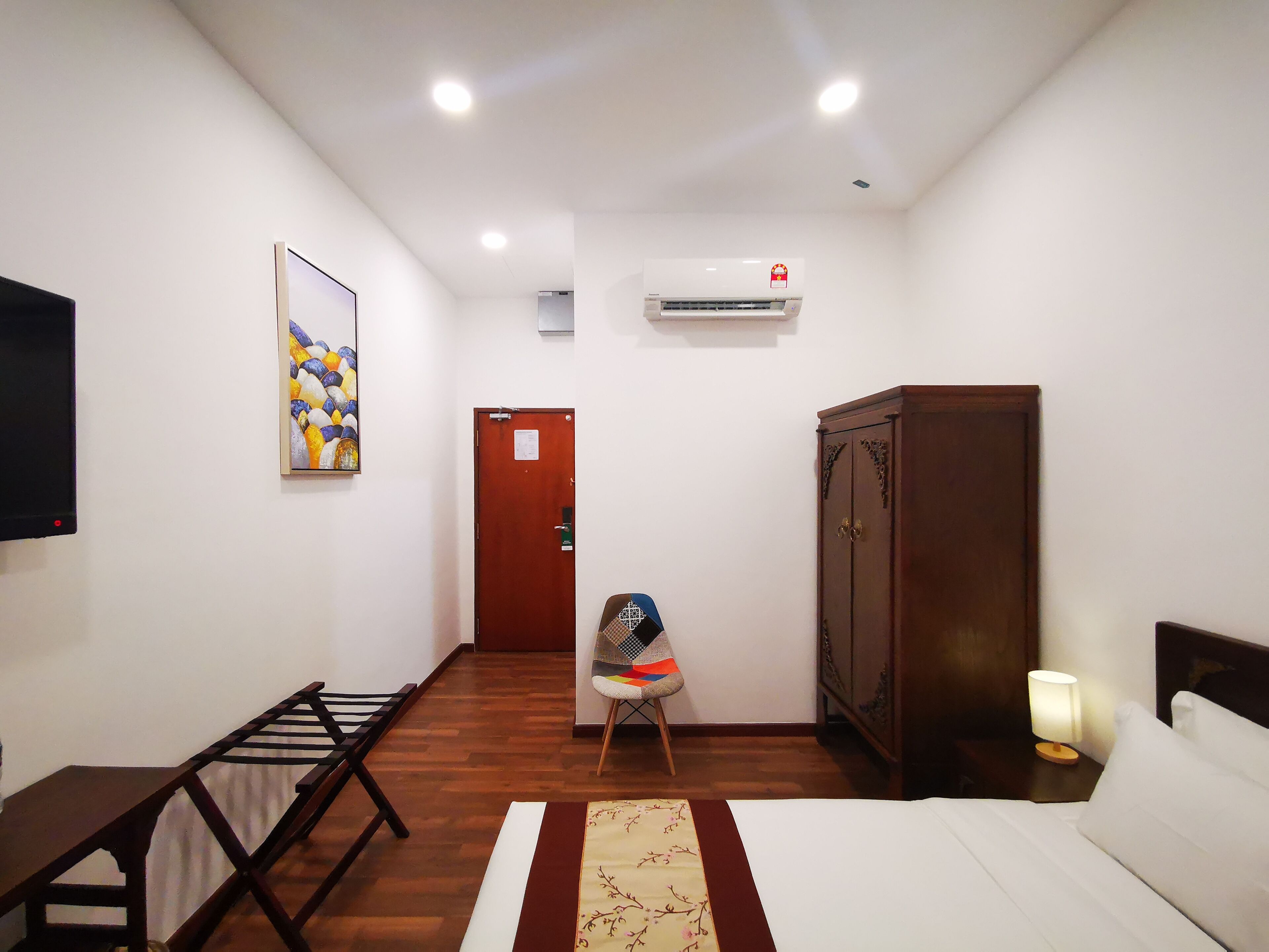 A hotel room with a bed, a wooden wardrobe, a desk with a lamp, a chair, a TV, an air conditioner, and a painting on the wall.