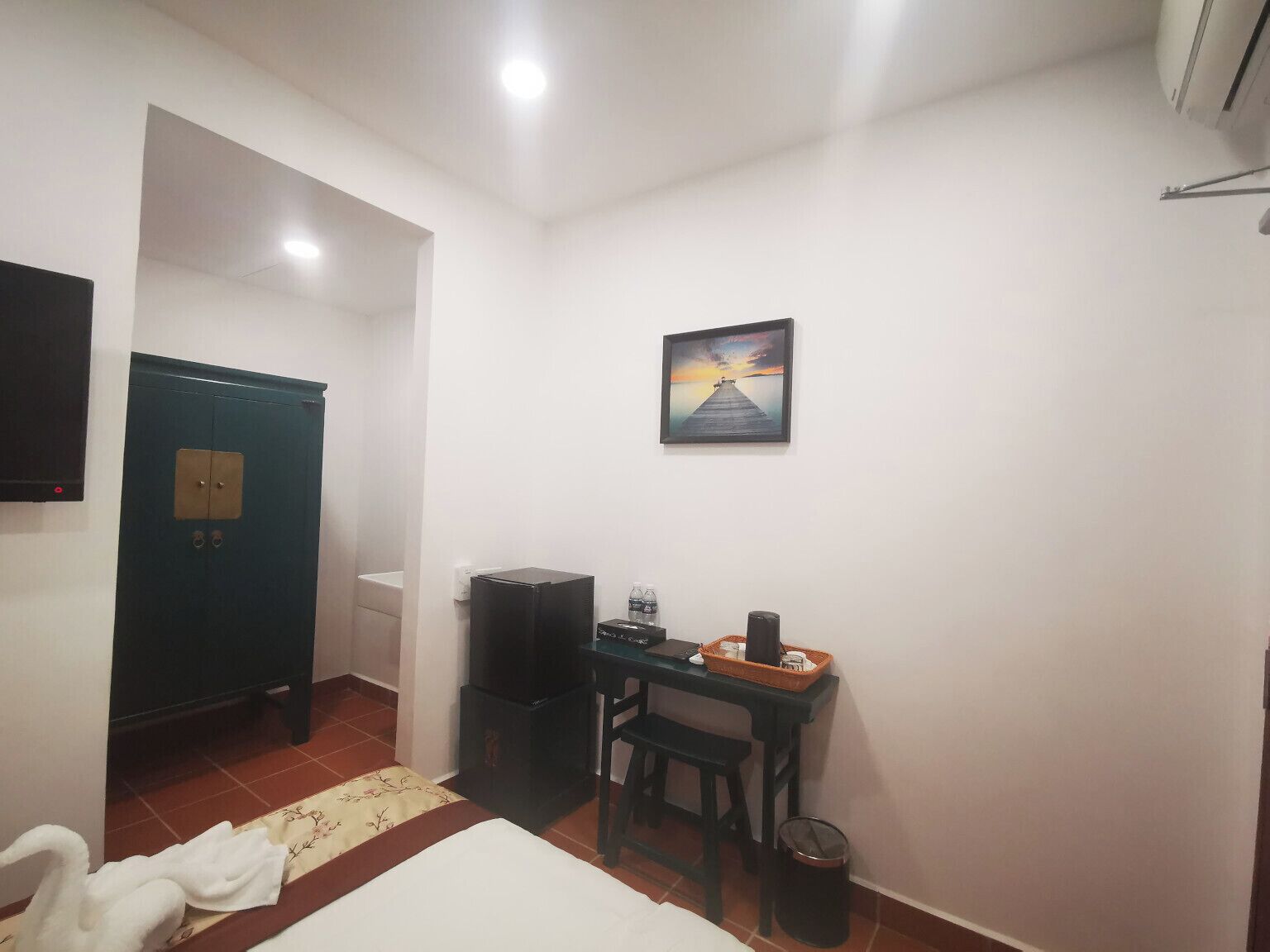 A room with a bed, a desk with a chair, a trash bin, a television, and a painting on the wall.