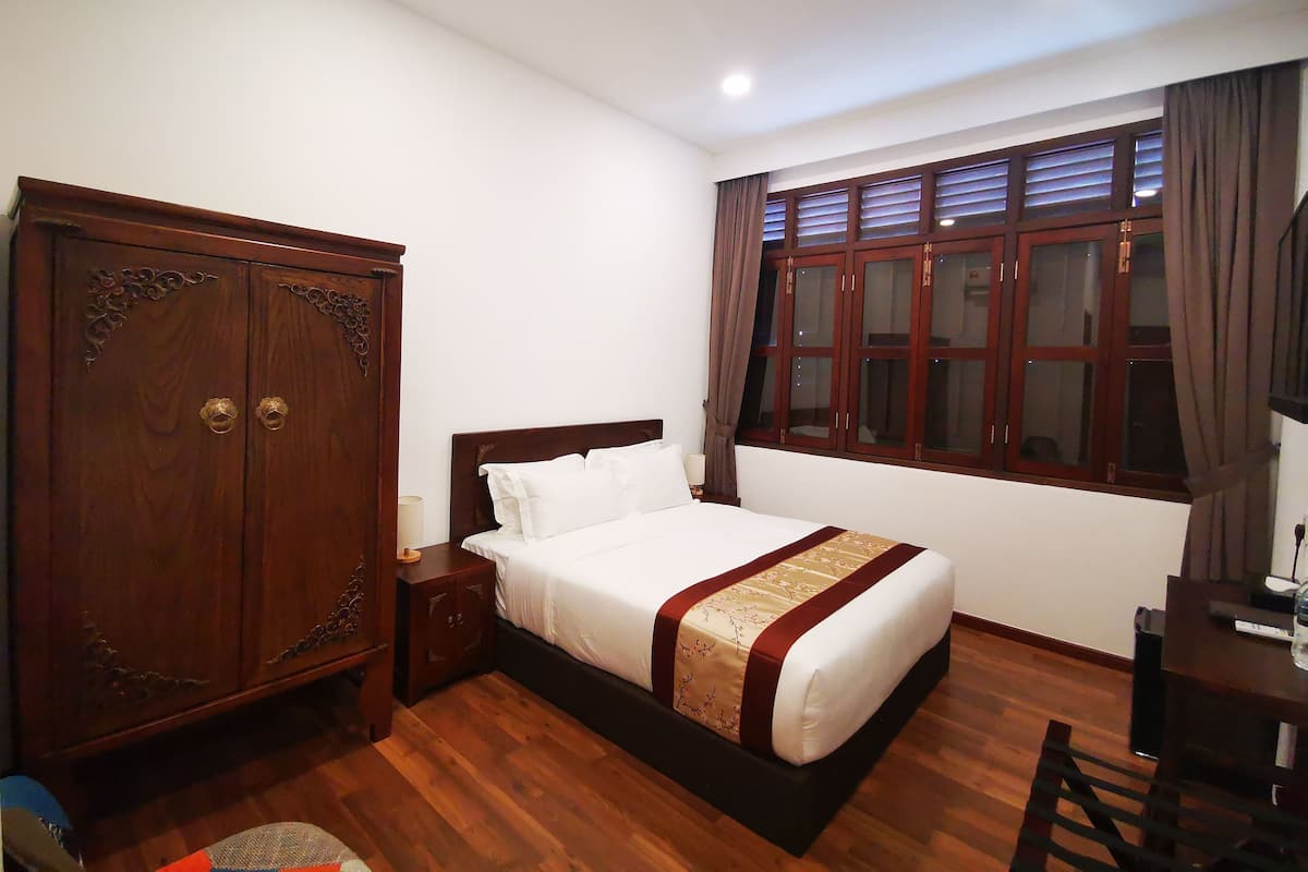 Ein Schlafzimmer mit einem Holzschrank, einem Bett mit weißer und roter Bettdecke, einem Nachttisch, einem Schreibtisch mit Stuhl und einem Fenster mit Vorhängen.