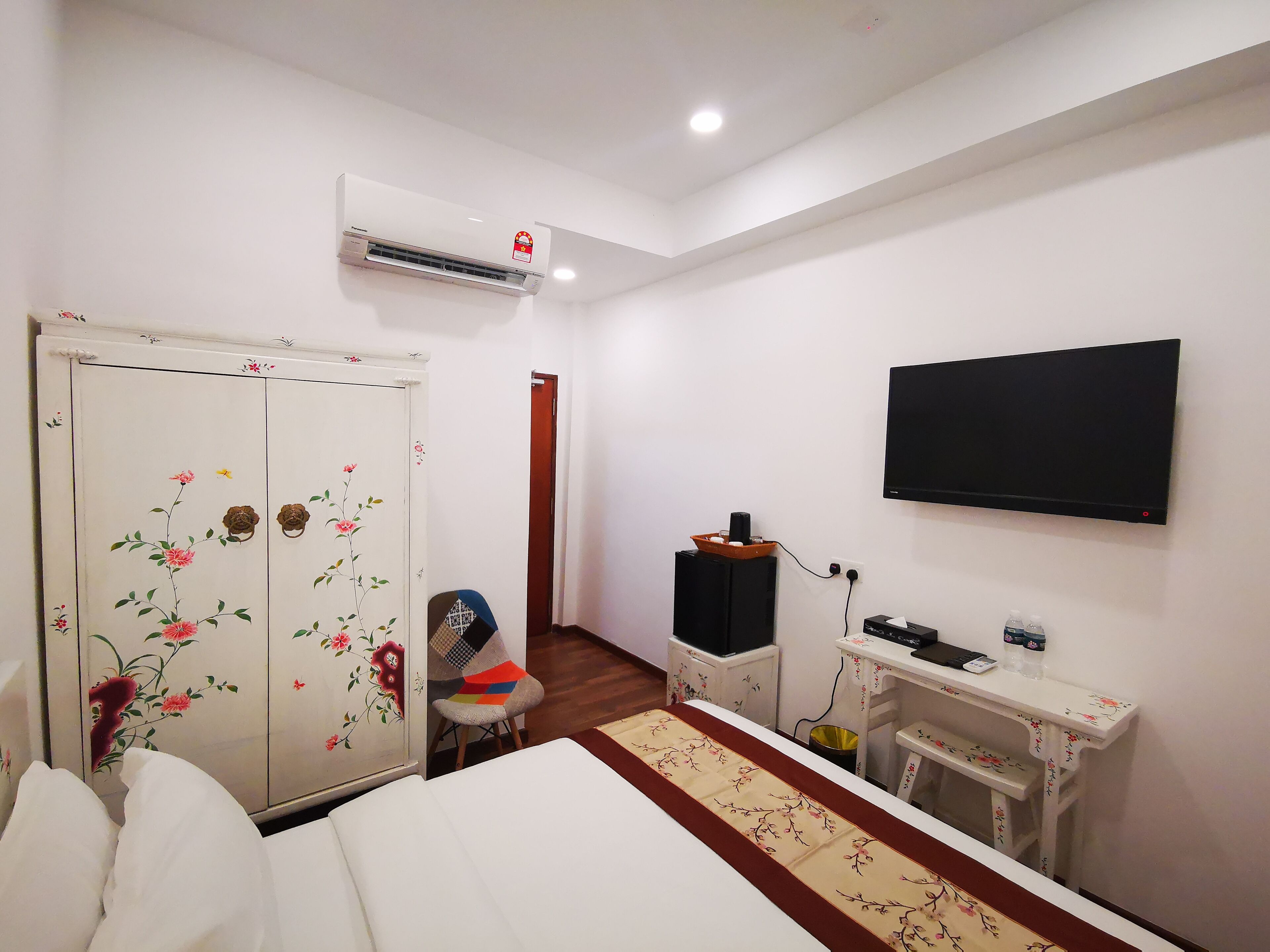 Une chambre d’hôtel comprenant un lit, une armoire ornée de motifs floraux, un bureau avec un ordinateur, une chaise et un téléviseur fixé au mur.