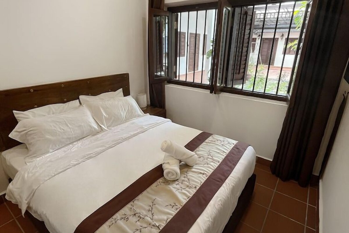 A neatly made bed with white bedding and a brown accent, a window with metal bars, and a view of the outside.