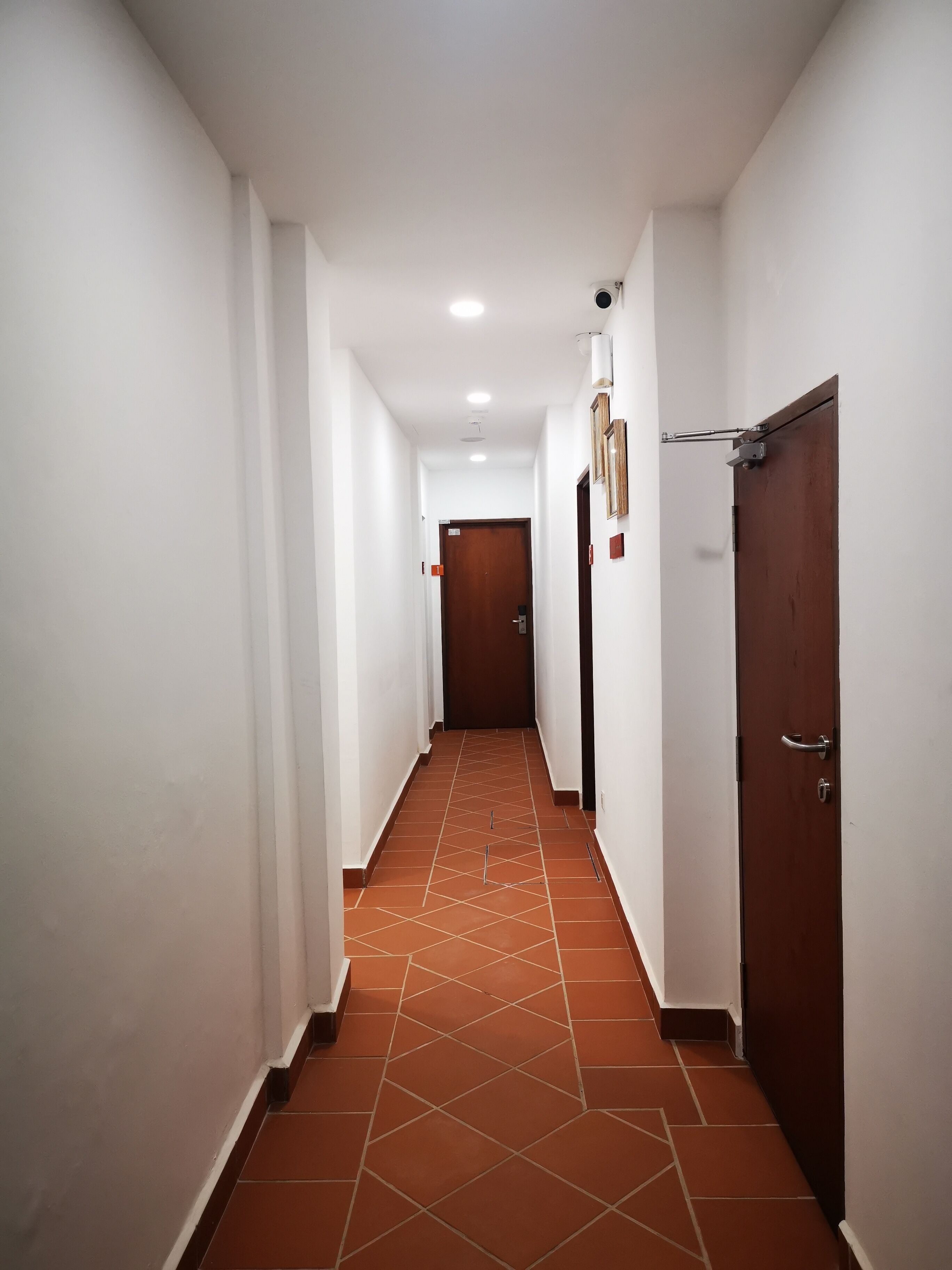 Un couloir aux murs blancs, une porte marron au fond et un sol carrelé.