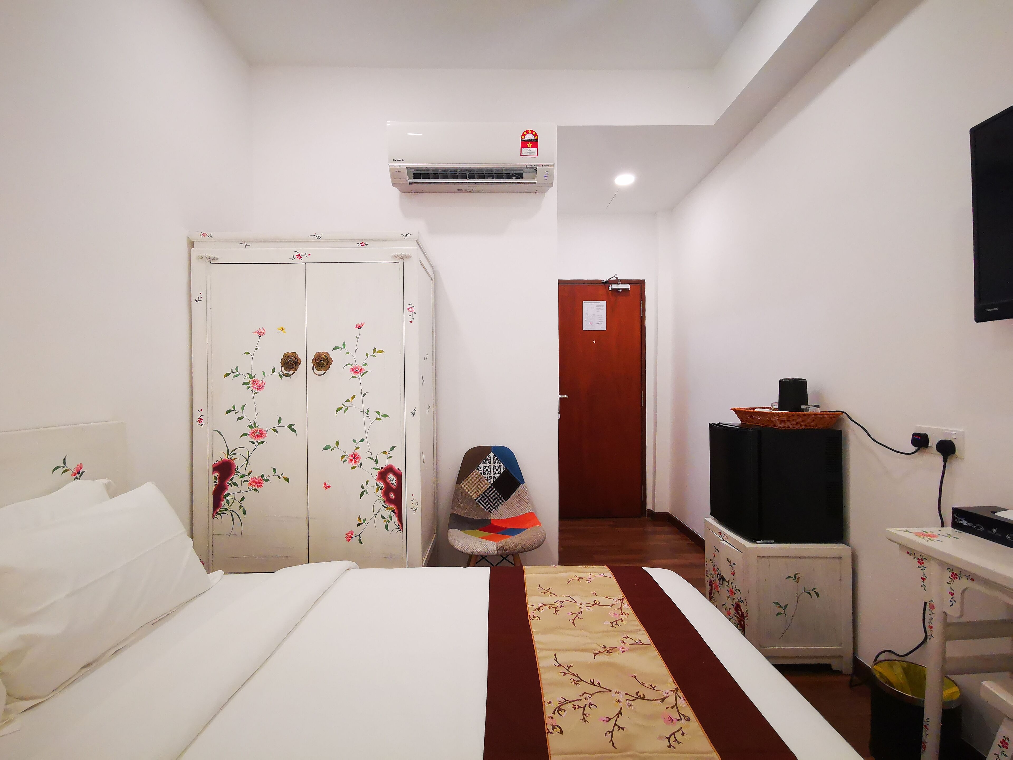 A hotel room with a bed, a wardrobe with floral designs, an air conditioning unit, a television, and a small table with a floral tablecloth.