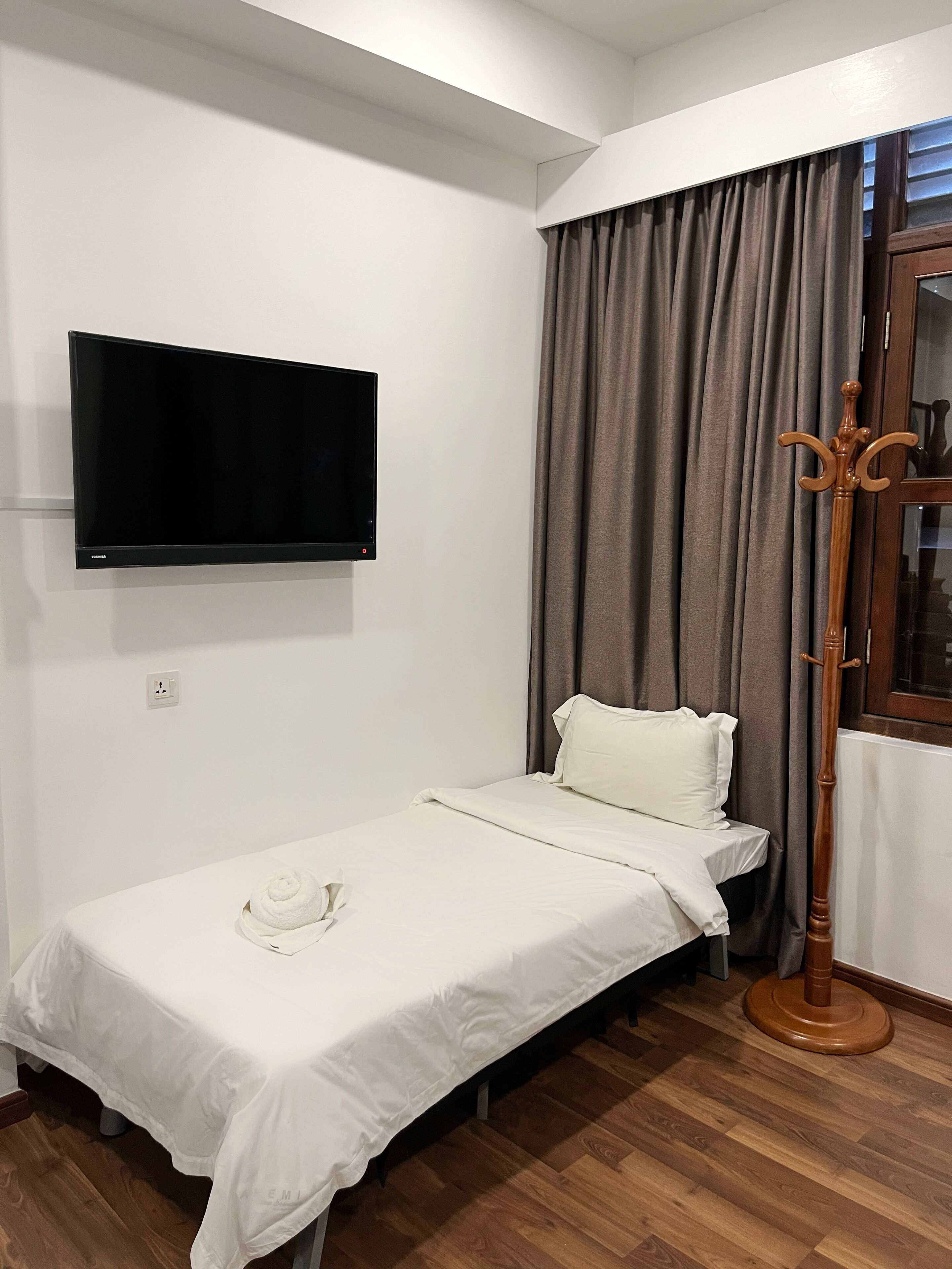 Un petit lit bien fait, avec un drap blanc, un téléviseur à écran plat fixé au mur et un porte-manteau en bois.