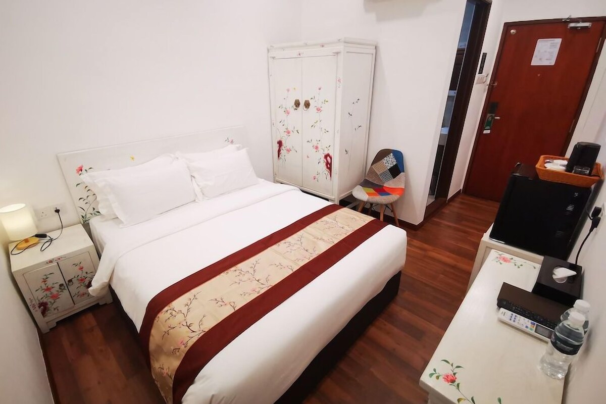 A hotel room with a bed, a wardrobe, a desk with a computer, a chair, and a bedside table with a lamp.