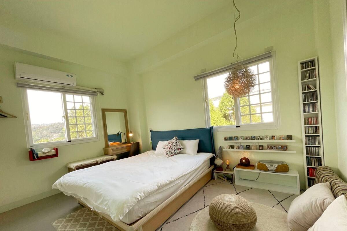 Een slaapkamer met een bed, nachtkastjes, een boekenkast en een raam met uitzicht op groen.