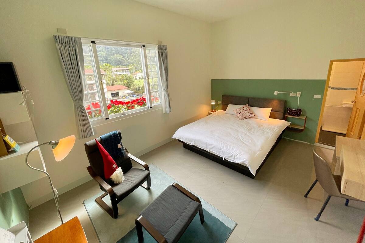 Een hotelkamer met een bed, een stoel, een bureau en een raam met uitzicht op gebouwen en bomen.