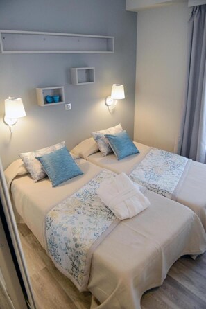 Ein Doppelbett mit floralem Bettbezug, zwei Kissen und ein gefaltetes Handtuch. Zwei Wandleuchten und ein kleines Regal befinden sich über dem Bett.