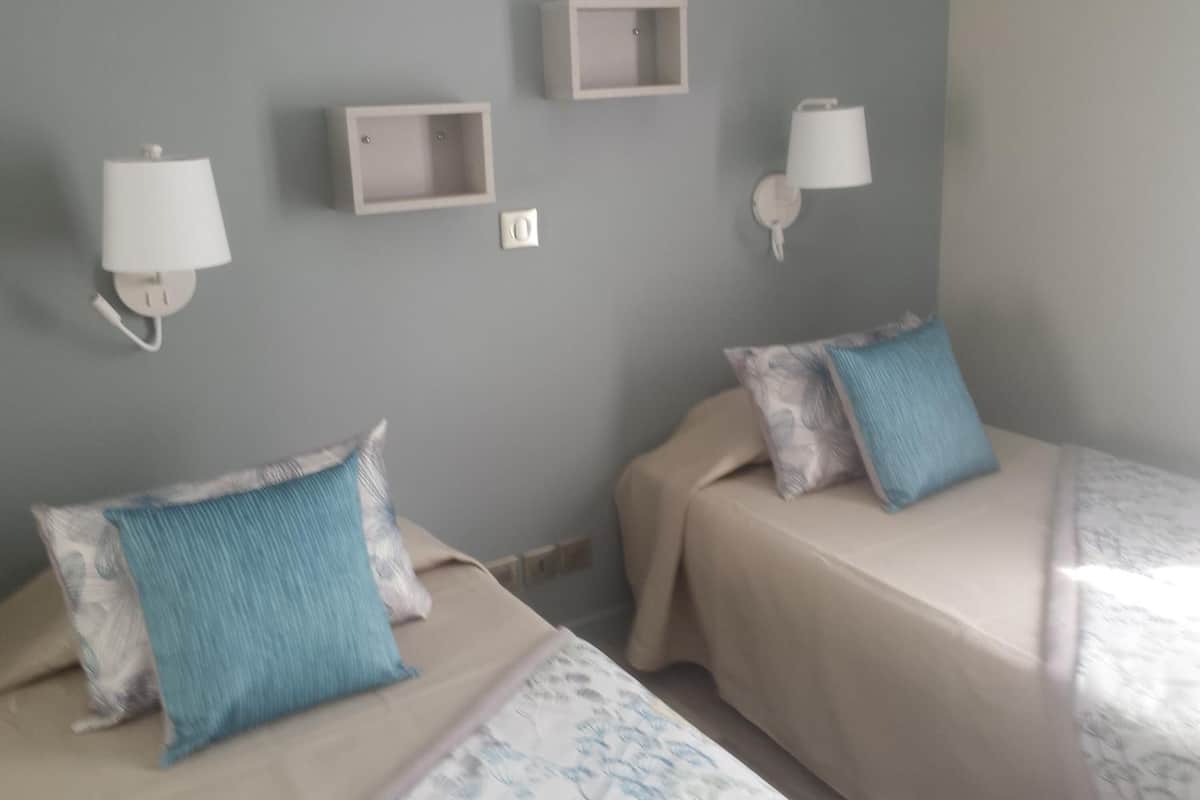 A room with two beds, each with a blue pillow and a white headboard. There are wall-mounted light fixtures and a small shelf above the beds.