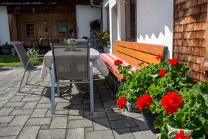 Une terrasse extérieure avec une table et des chaises, un banc en bois et des pots de fleurs rouges.
