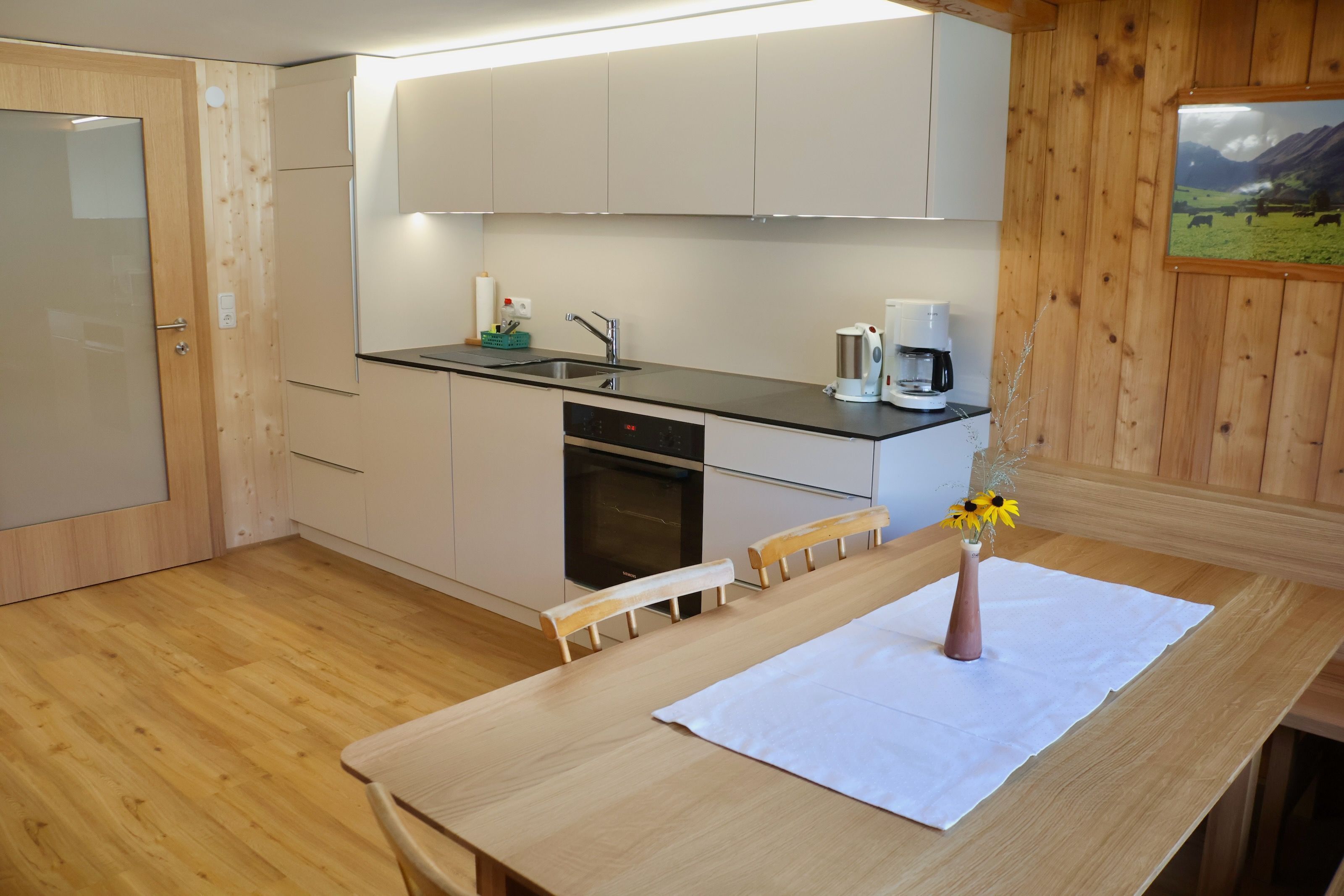 Une cuisine moderne avec des meubles en bois, un four encastré, une machine à café et une table à manger agrémentée d’un vase de fleurs.