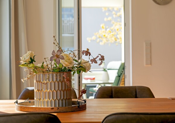 Une table à manger ornée d’un vase de fleurs, d’un plateau en bois, et une fenêtre donnant sur un bâtiment et des arbres.