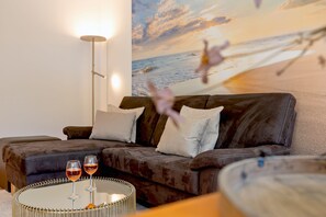 A living room with a brown leather sofa, two white pillows, a glass coffee table with two wine glasses, and a wall mural of a beach scene.