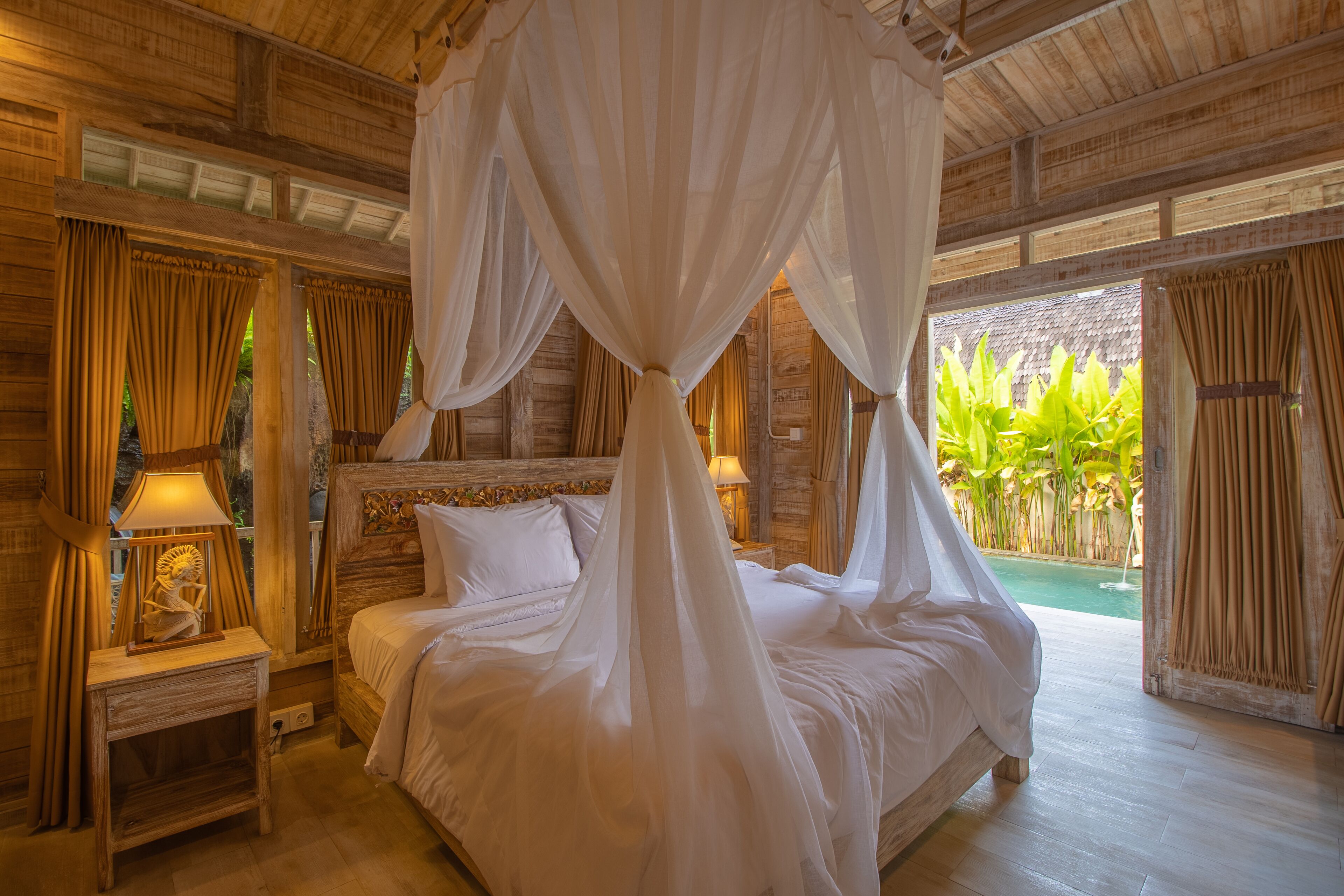 A room with a bed under a canopy, wooden walls, a nightstand with a lamp, and a view of greenery outside.