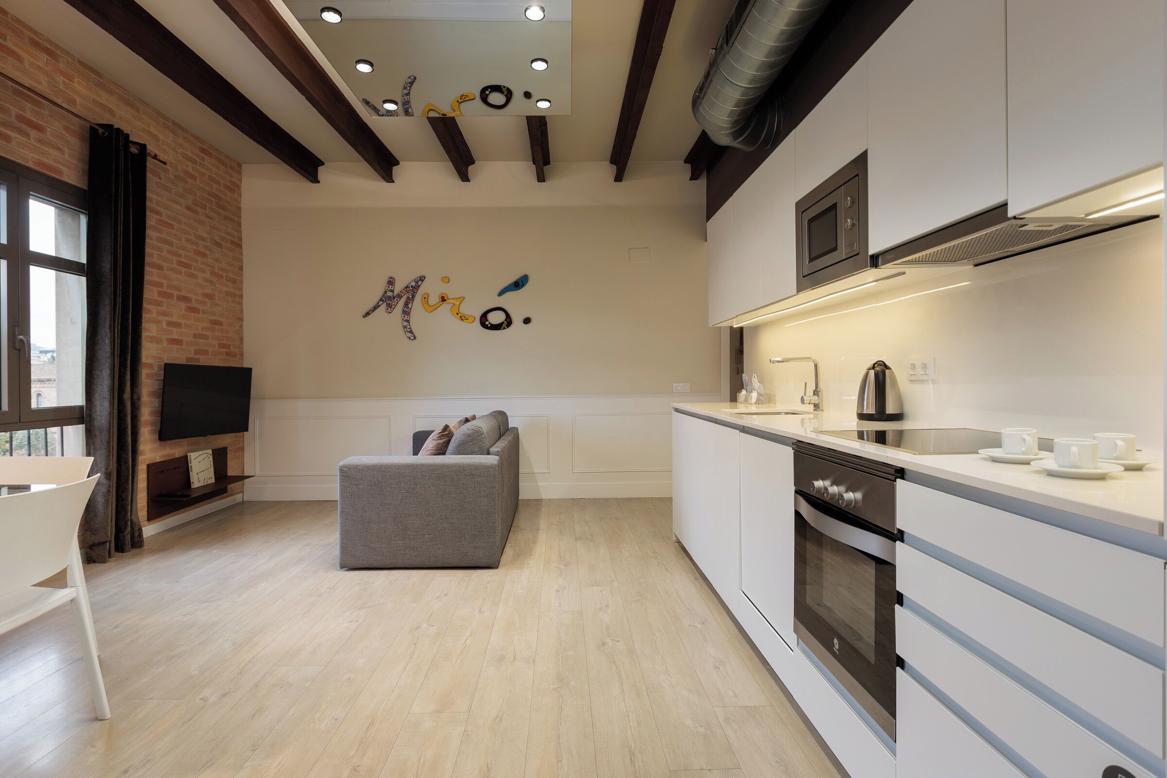 A modern kitchen with white cabinets, a built-in oven, and a microwave. There is a grey sofa and a flat-screen TV mounted on the wall.